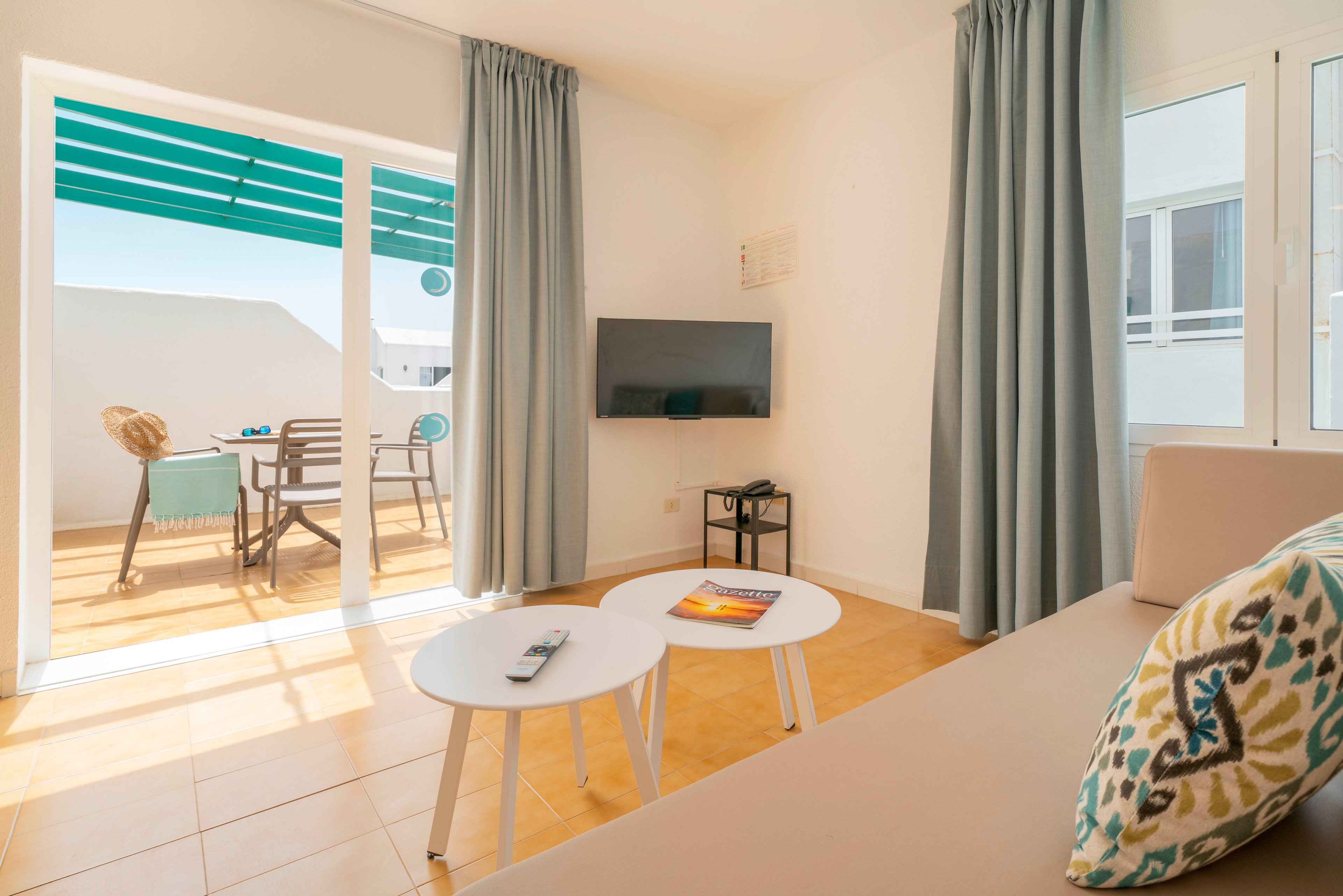 Sunlight fills a modern apartment living room with a sofa, nesting coffee tables, and a wall-mounted television. Large glass doors open onto a sunny terrace with a table, chairs, and a green pergola. Soft blue curtains and neutral furnishings create a calm, coastal atmosphere. The space appears tidy and inviting, ideal for a holiday rental or short stay.