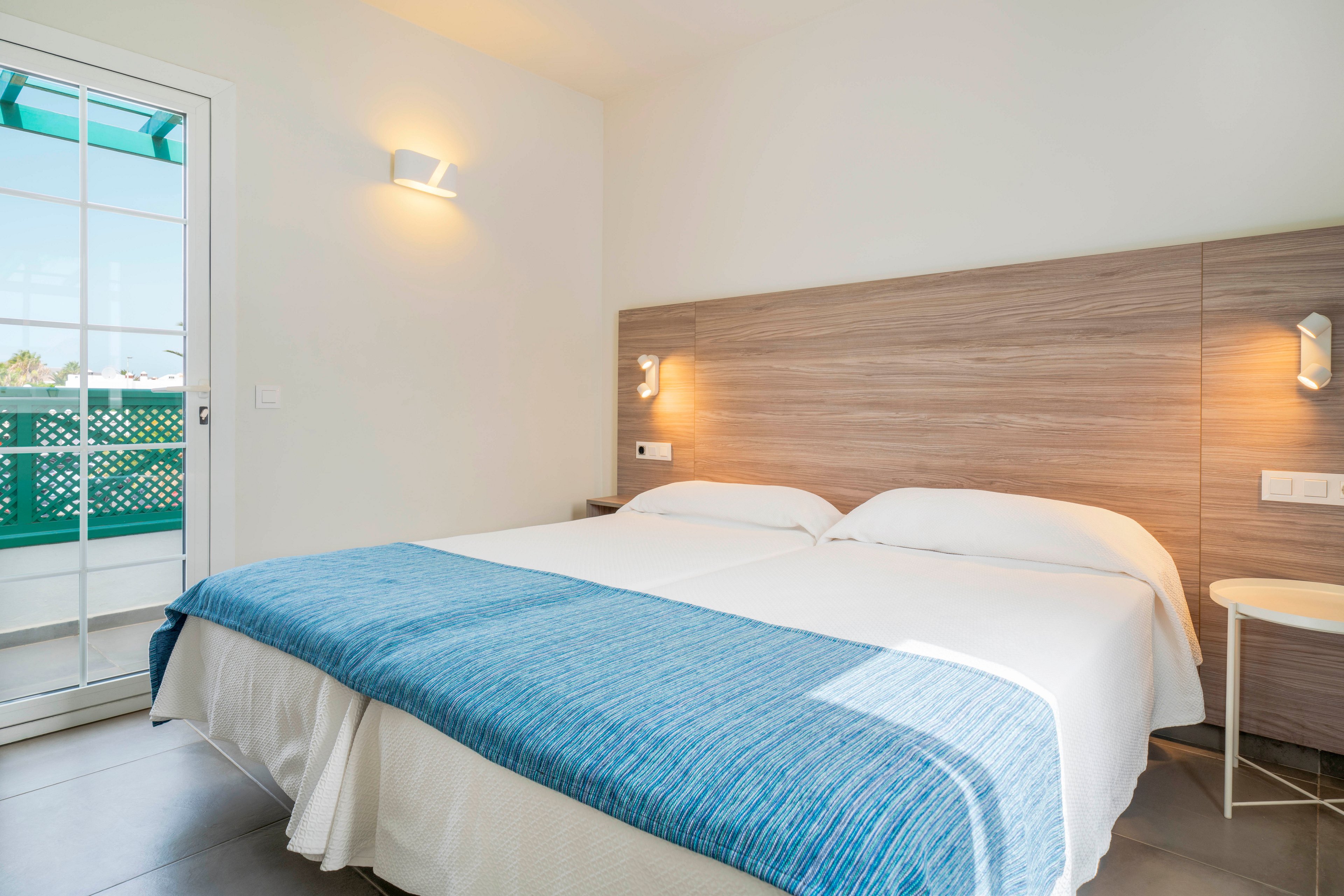 A clean, modern hotel bedroom features two single beds pushed together with white linens and a blue throw blanket. The room has a light wood headboard with built-in reading lights and a small bedside table. Natural light enters through a glass door leading to a balcony with green lattice panels. The overall space feels minimal, airy, and inviting.