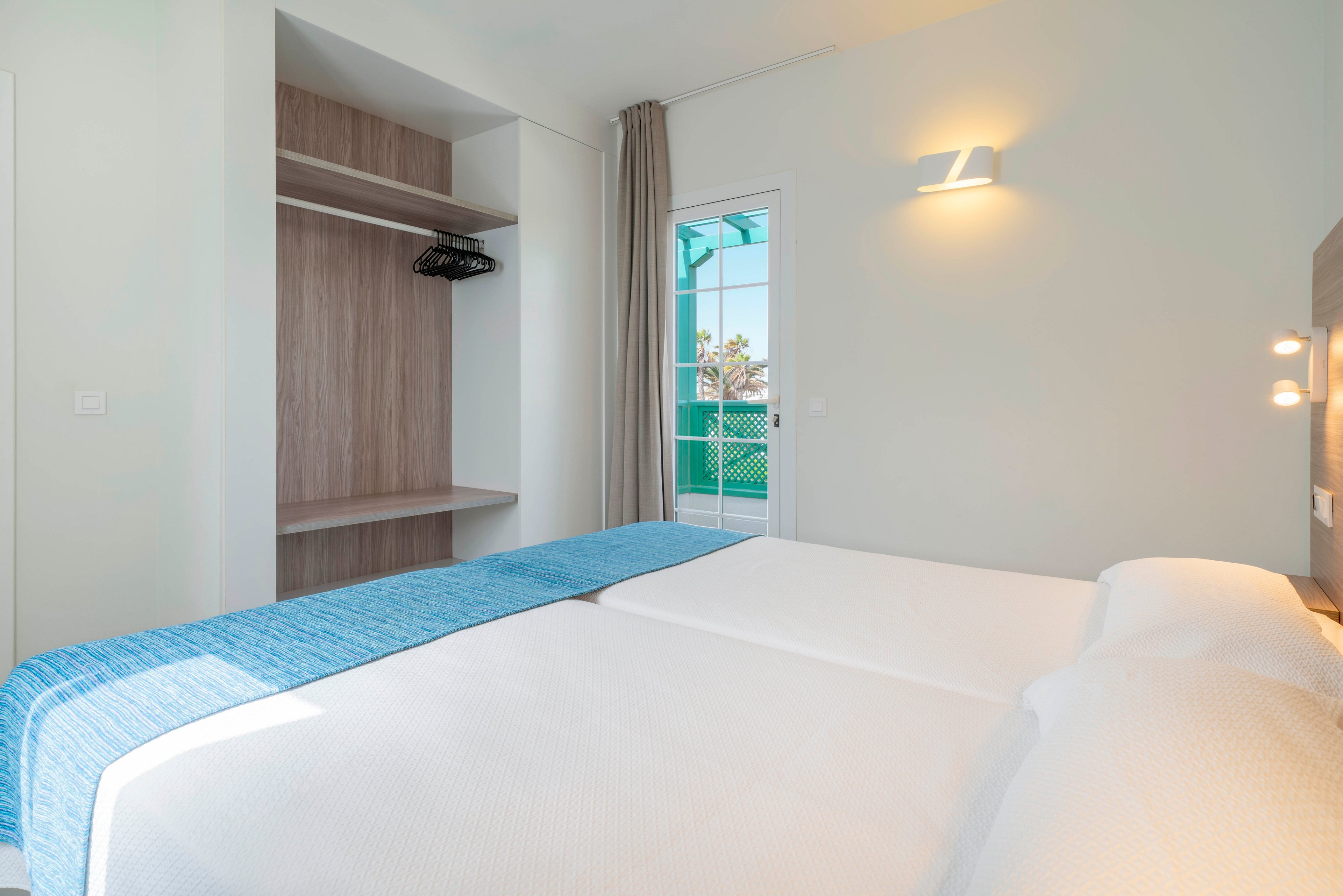 A bright, minimalist hotel bedroom features two neatly made single beds pushed together with white linens and a blue runner. An open wooden wardrobe with shelves and hangers stands to the left, adding functional storage space. Natural light enters through a glass door leading to a balcony with green railings and a partial outdoor view. Soft wall lighting and neutral walls create a calm, contemporary atmosphere suitable for relaxation.
