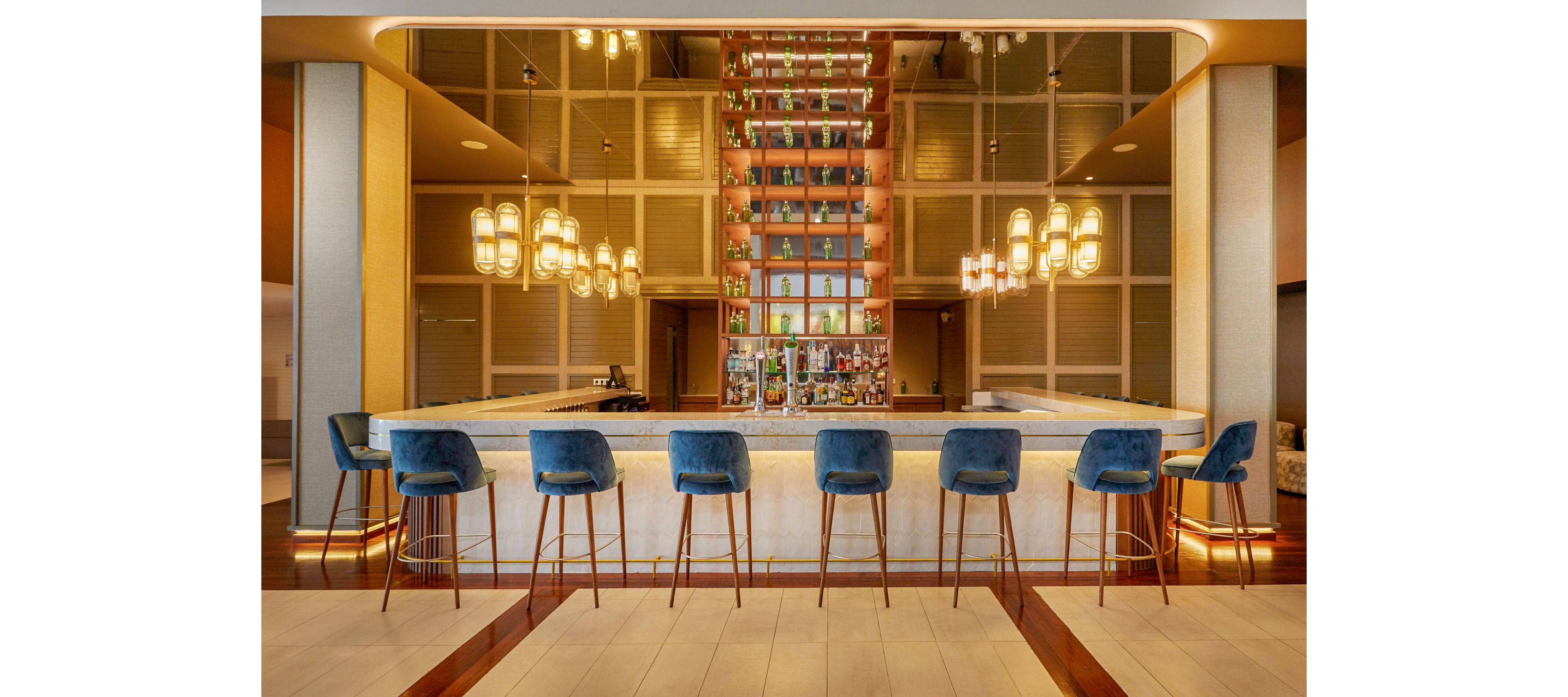 A contemporary hotel bar features a sleek marble counter surrounded by blue velvet bar stools. Warm pendant lights hang above the bar, casting a golden glow over the seating area. A tall mirrored back wall showcases neatly arranged bottles and glassware, adding depth and symmetry to the scene. The overall atmosphere feels upscale, inviting, and designed for relaxed socializing.