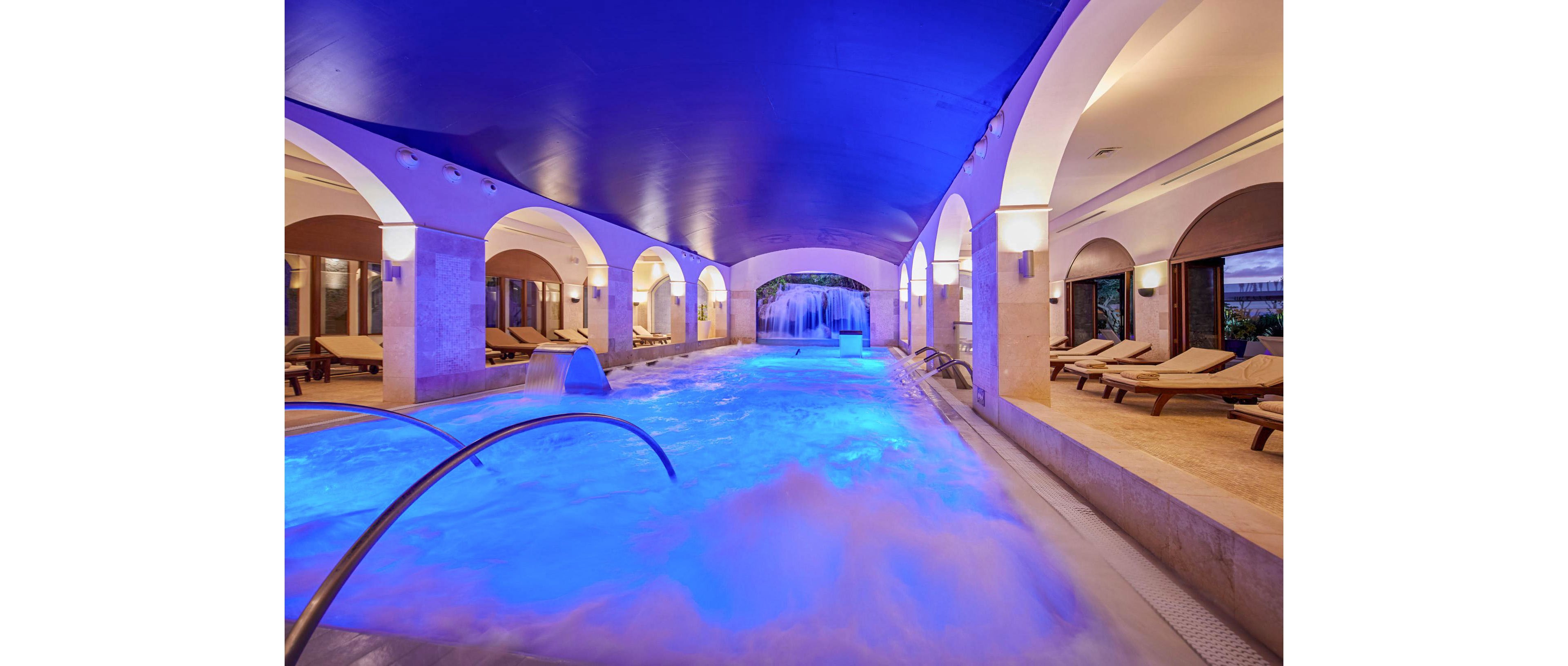 An elegant indoor spa pool glows with vibrant blue lighting beneath a smooth arched ceiling. Stone columns and arches frame the water, creating a refined and tranquil atmosphere. Multiple lounge chairs line the sides of the pool, inviting guests to relax. Water jets and a cascading waterfall add movement and a sense of luxury to the serene wellness setting.