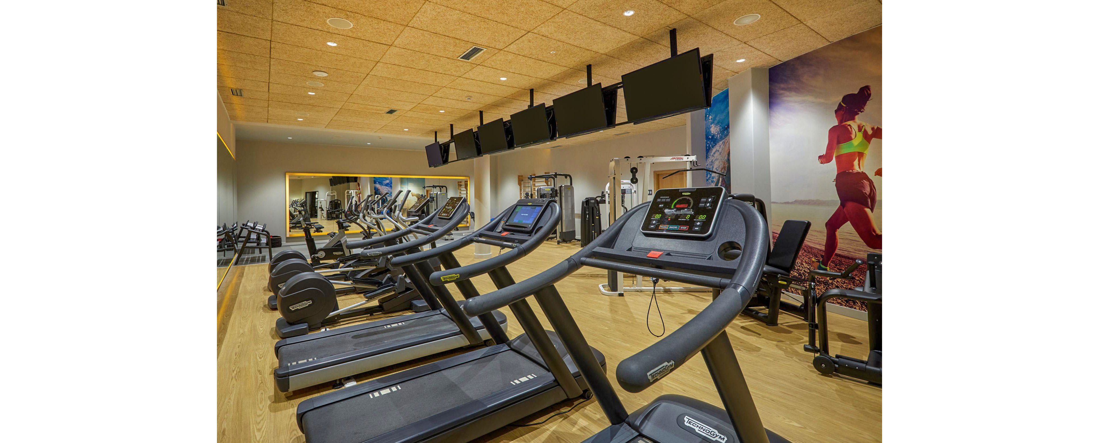 A modern fitness center features a row of treadmills lined up in the foreground. Large wall-mounted screens hang above the machines, facing users for entertainment or workout data. Additional strength and cardio equipment is visible in the background, reflected in a large wall mirror. Warm lighting, wooden flooring, and a colorful mural of a runner create an energetic yet welcoming atmosphere.