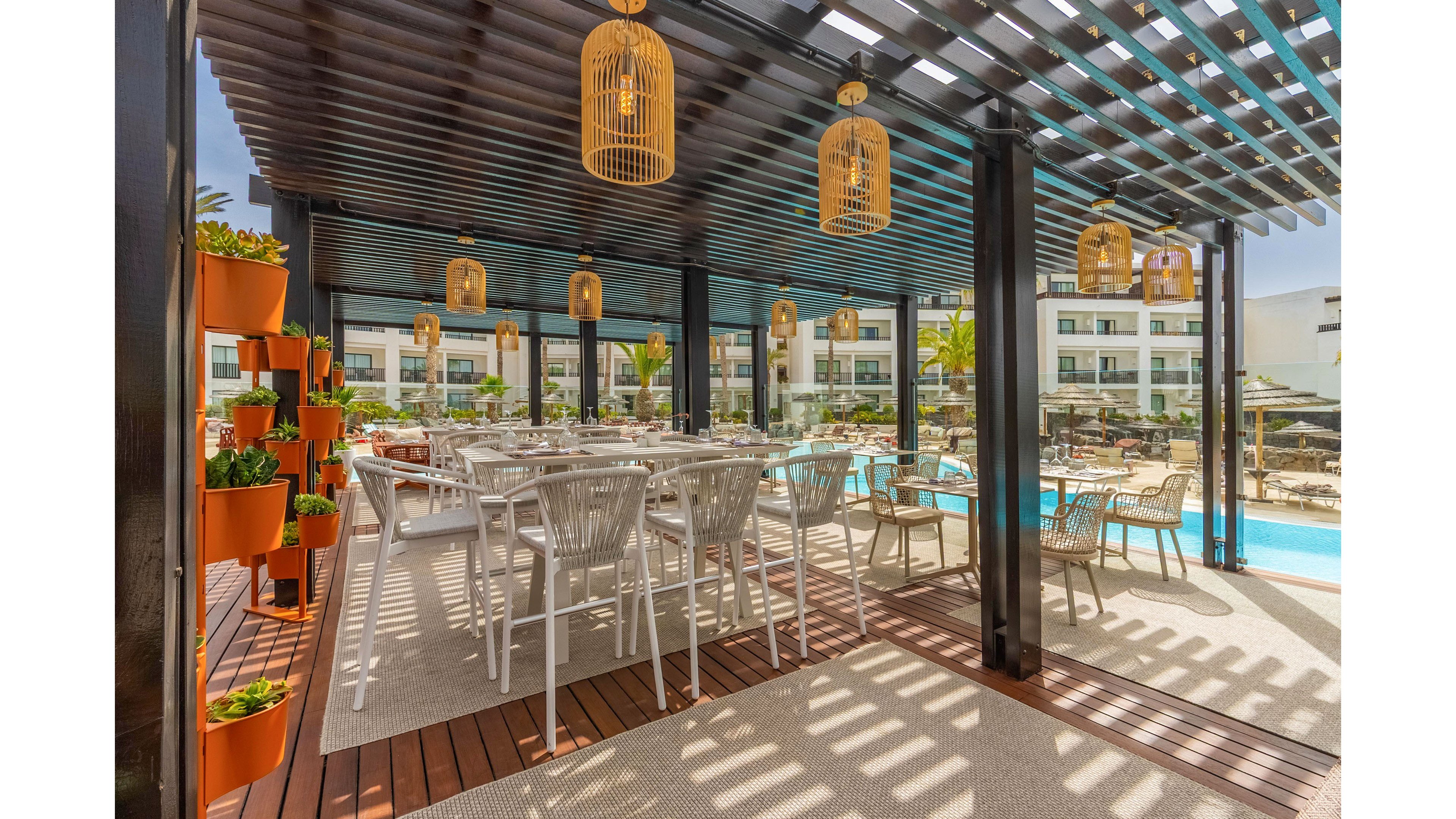 An open-air dining terrace is set beneath a modern pergola beside a bright blue swimming pool. White tables and chairs are arranged neatly, creating a relaxed yet stylish seating area. Decorative hanging lanterns and a vertical garden of orange planters add warmth and greenery to the scene. In the background, a contemporary hotel or resort building and sun loungers suggest a vacation or leisure setting.