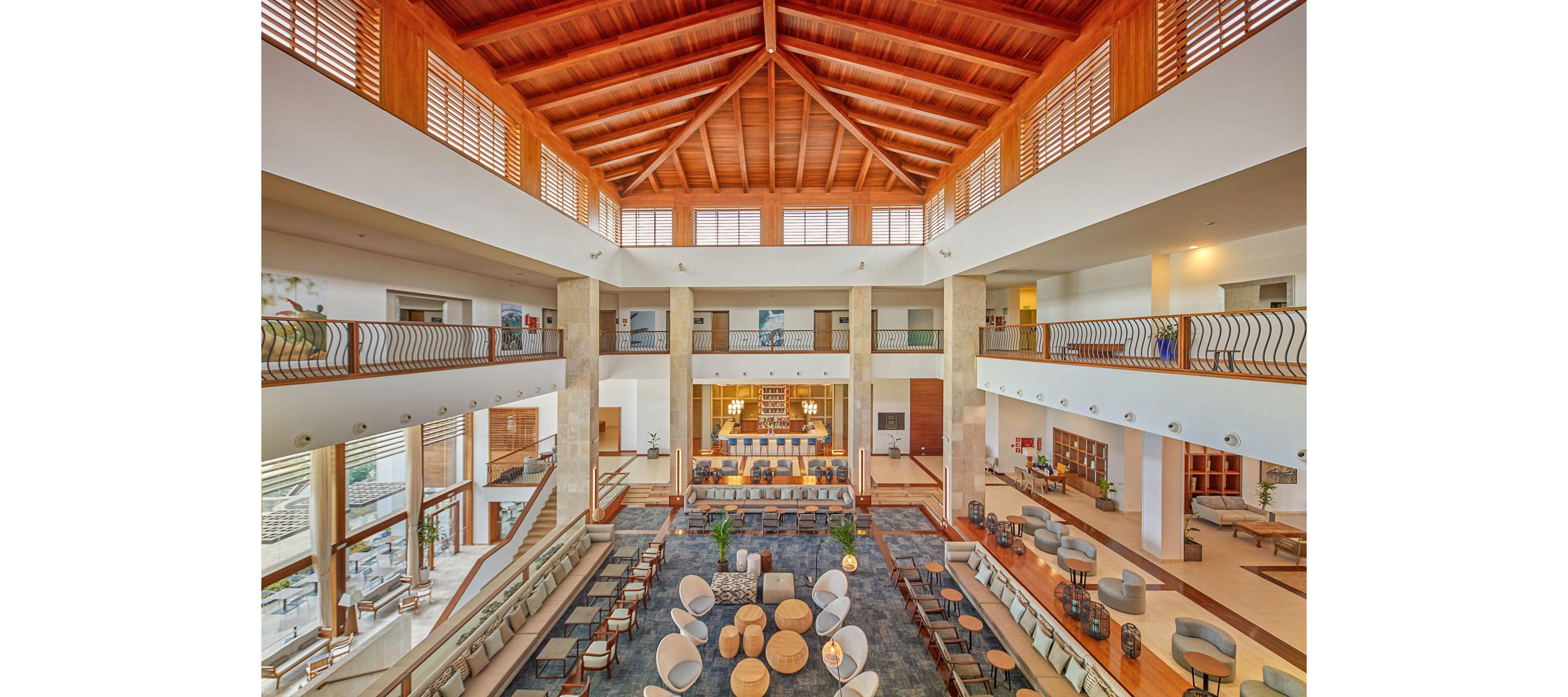 A large, contemporary hotel lobby features an open atrium with multiple levels and a high wooden ceiling. Comfortable seating areas with sofas, armchairs, and round tables are arranged symmetrically around a central rug. Warm natural light streams in through tall windows, highlighting the neutral tones and wooden accents. A bar or reception area is visible at the far end, creating an inviting social and relaxation space.