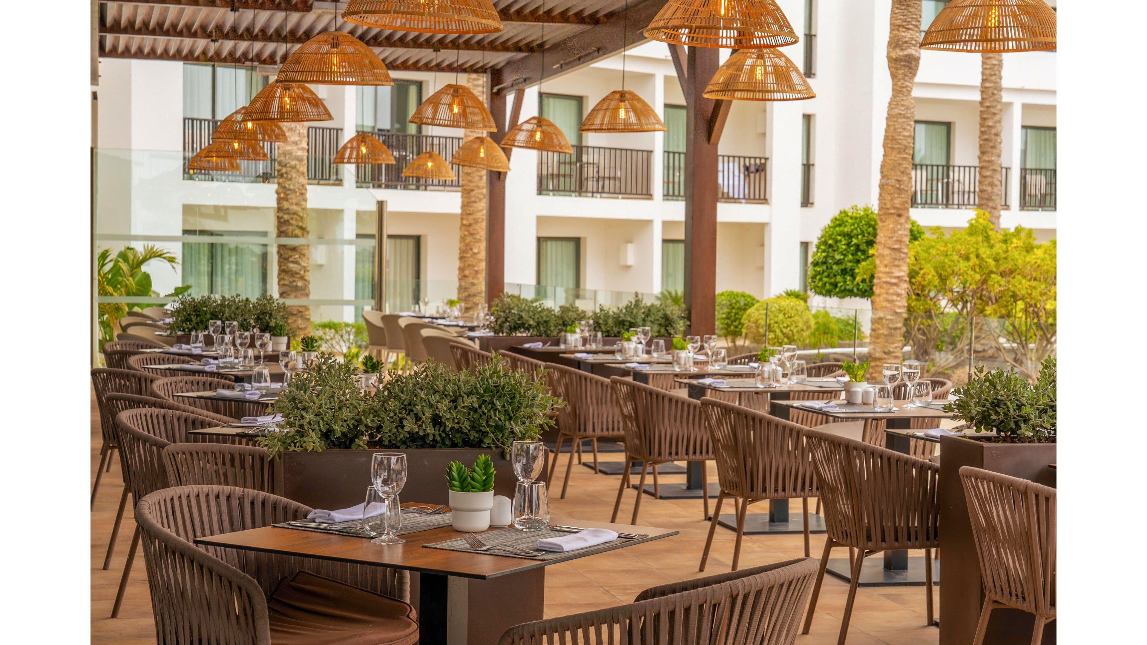 A contemporary outdoor restaurant terrace is set with neatly arranged tables and chairs. Each table is prepared with glassware, cutlery, and placemats, ready for guests. Warm pendant lights hang from the wooden pergola above, creating a cozy ambiance. In the background, a modern hotel building, balconies, and palm trees frame the relaxed resort setting.