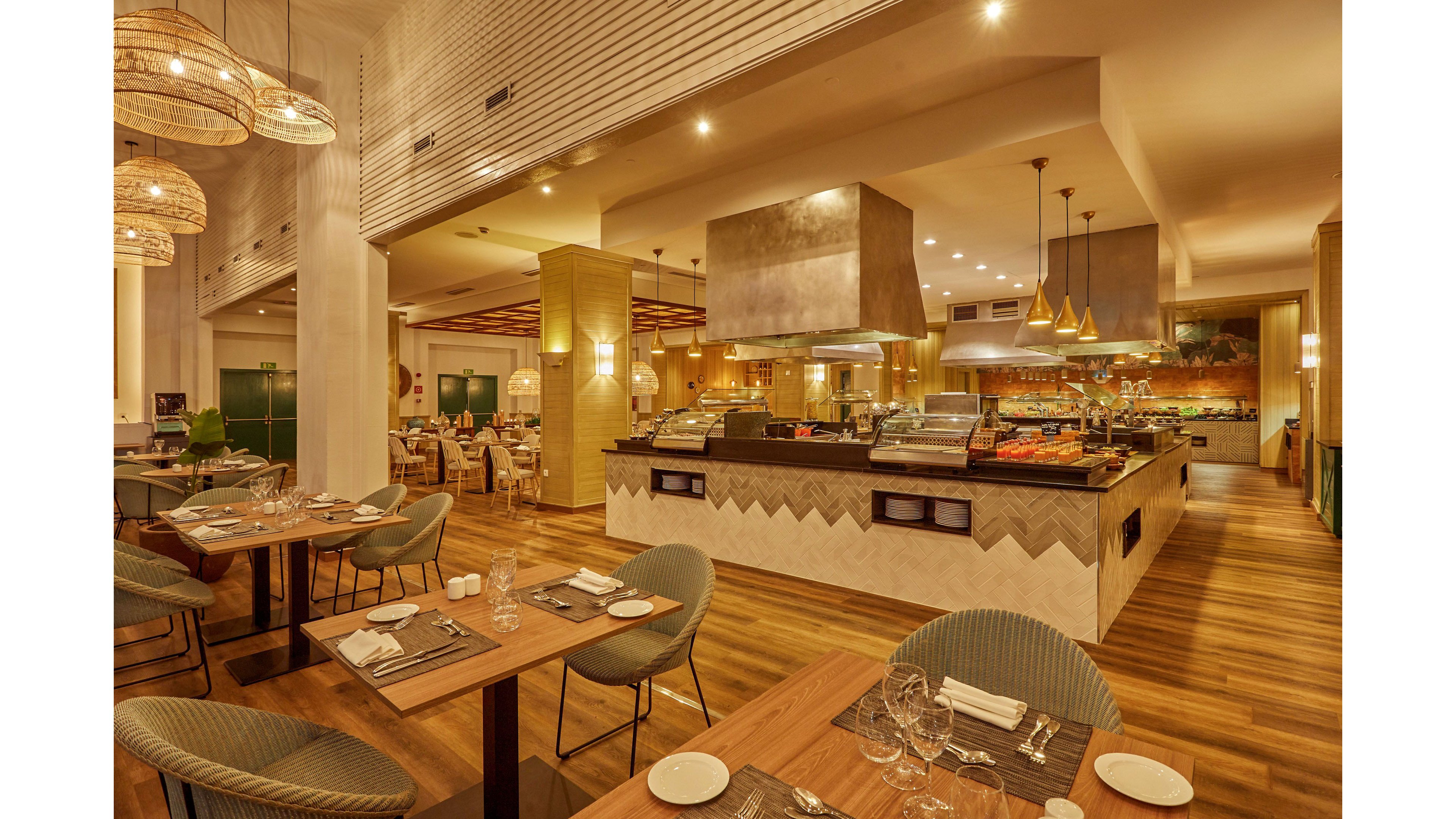 A contemporary restaurant dining area features neatly set wooden tables and upholstered chairs. In the center, a large buffet island displays covered dishes and serving stations under metallic hoods. Warm pendant lights and woven lamps create a cozy, golden ambiance across the space. The wooden flooring and neutral-toned decor contribute to an inviting, upscale atmosphere. No guests are present, highlighting the clean and orderly setup.
