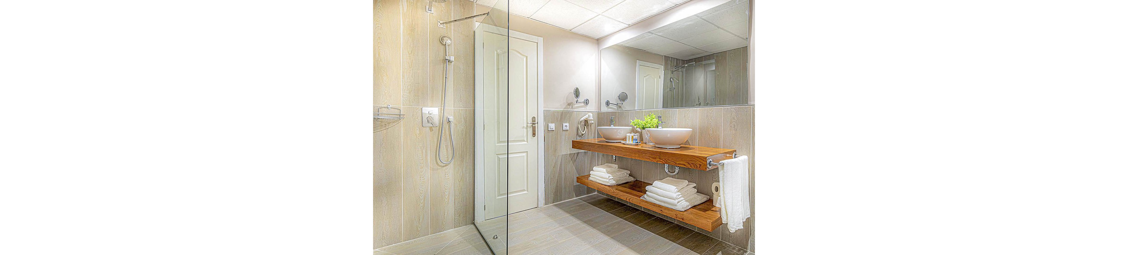 A contemporary bathroom featuring a wooden vanity with two vessel sinks and neatly folded towels. The space includes a glass-enclosed shower and a white door, with beige tiled walls and flooring. A small green plant adds a touch of freshness to the clean and minimalist design.