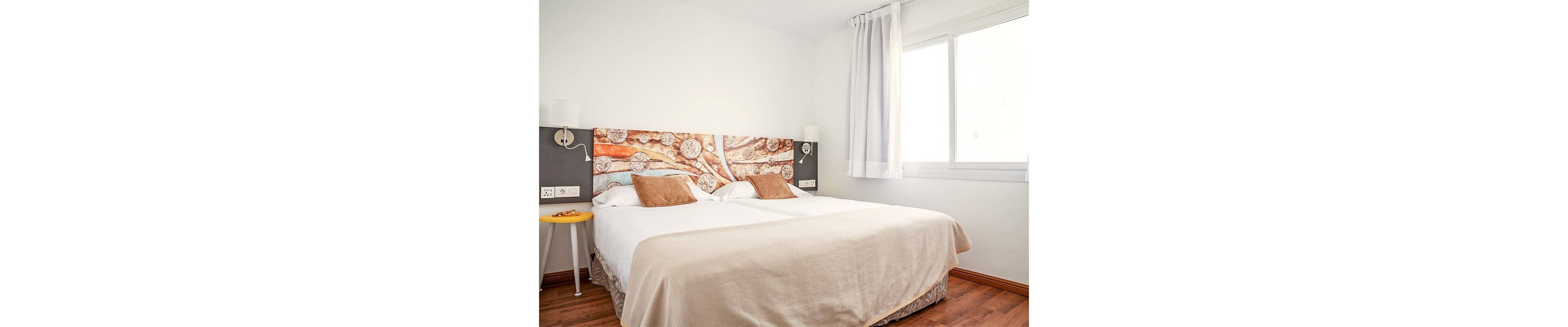 A bright, minimalist bedroom features a large double bed with a decorative headboard displaying abstract artwork. Neutral bedding in beige and white is accented by several brown throw pillows. The room includes wall-mounted reading lamps, a small yellow side table, and a wooden floor. Natural light streams in through a large window with white curtains, creating a clean and inviting atmosphere.