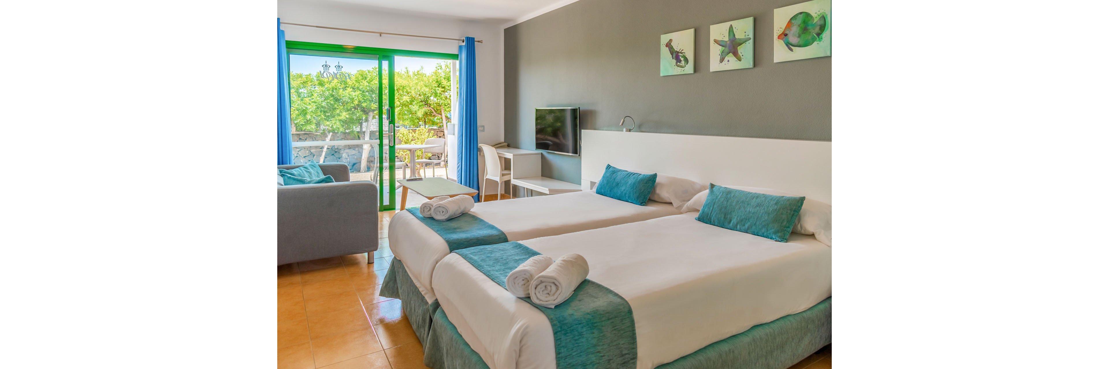 A modern hotel room features two neatly made twin beds with white linens and turquoise accents, each topped with rolled towels. The space includes a wall-mounted flat-screen TV, a small desk and chair, and a gray sofa. Large glass doors with bright blue curtains open to a balcony overlooking lush green trees, filling the room with natural light. The decor is simple and coastal, with three small artworks above the beds and a clean, inviting atmosphere.
