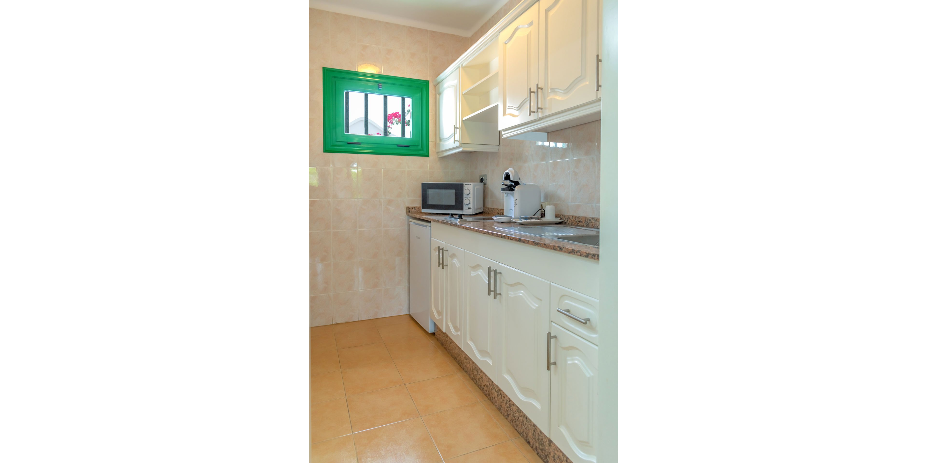 A compact, narrow kitchen features white cabinets and a granite-style countertop along one wall. A microwave and a small countertop appliance sit beside a stainless steel sink. Beige tiled walls and floors create a warm, neutral backdrop. A green-framed window brings in natural light and adds a pop of color to the space.