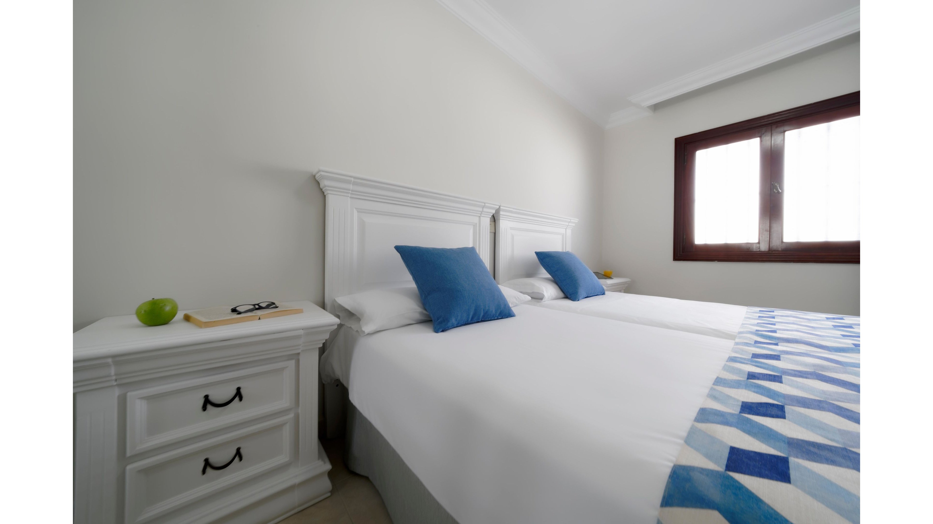 A clean, minimalist bedroom features two neatly made twin beds with white linens and blue accent pillows. A geometric blue and white bed runner adds subtle pattern and color to the scene. White bedside tables flank the beds, one holding an apple and a pair of eyeglasses. Natural light streams in through a wooden-framed window, creating a calm and inviting atmosphere.