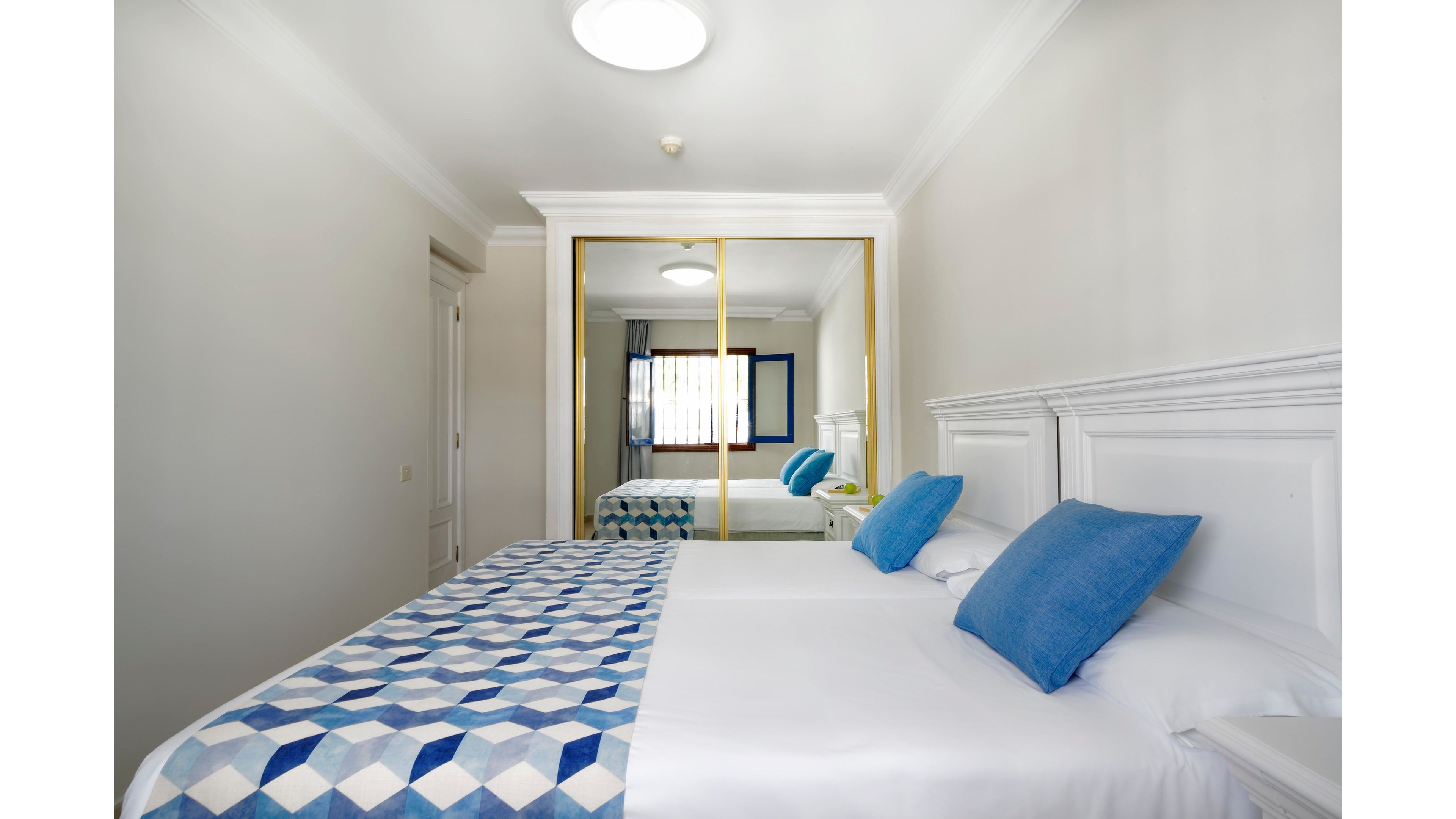 A clean, modern bedroom features two neatly made twin beds with white linens and geometric blue patterned runners. Blue throw pillows add color against the white headboards and walls. A large mirrored wardrobe reflects the window, enhancing the sense of space and light. The room has a minimalist, tidy feel with soft natural and ceiling lighting.