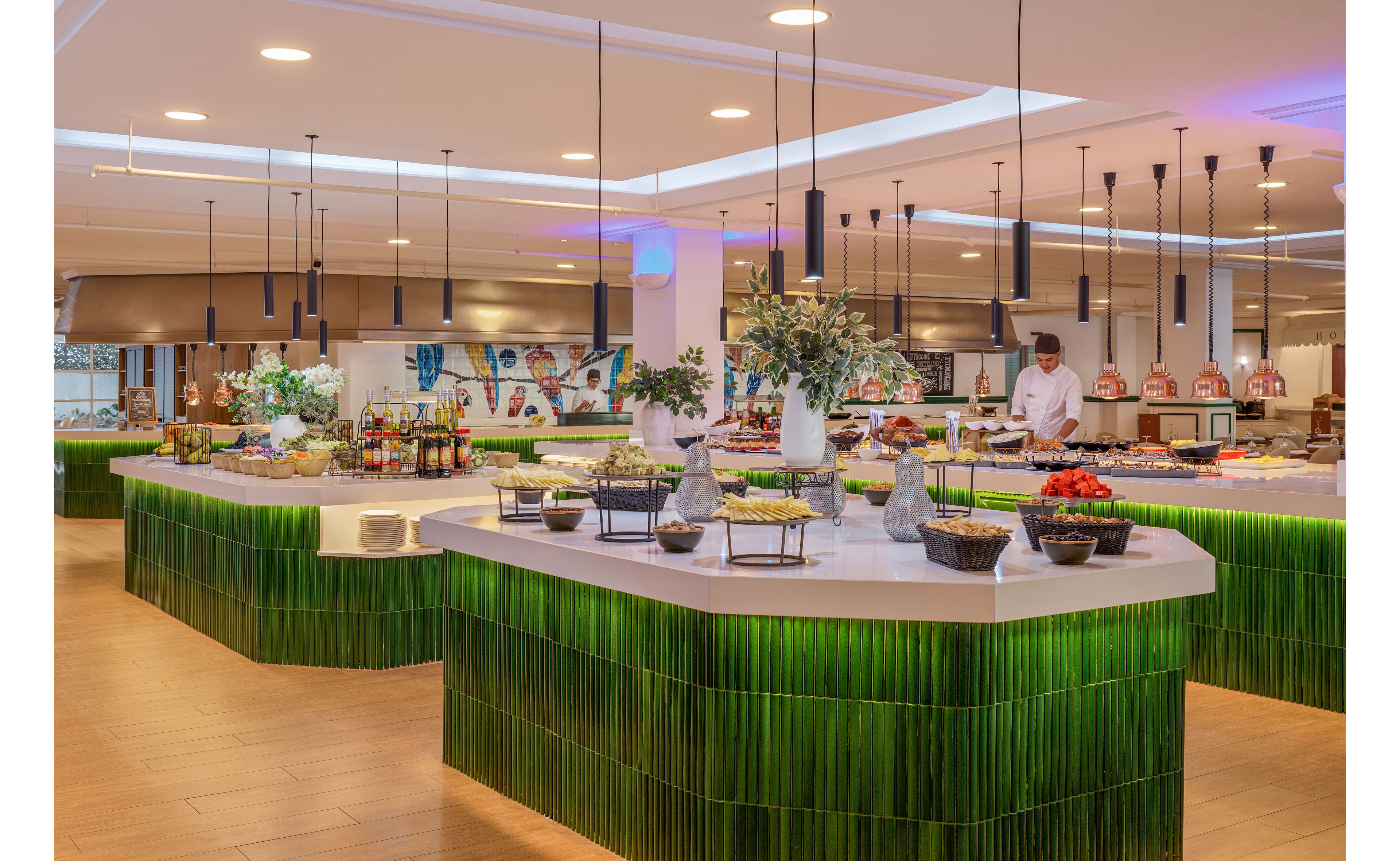 A spacious, contemporary buffet area features long counters with green textured panels and neatly arranged food displays. Bowls of salads, fruits, and other dishes are set out under warm ceiling lights, creating an inviting dining atmosphere. A chef in a white uniform and hat works behind the counters, with ovens and cooking stations visible in the background. Decorative plants and wall art add color and style to the bright interior.