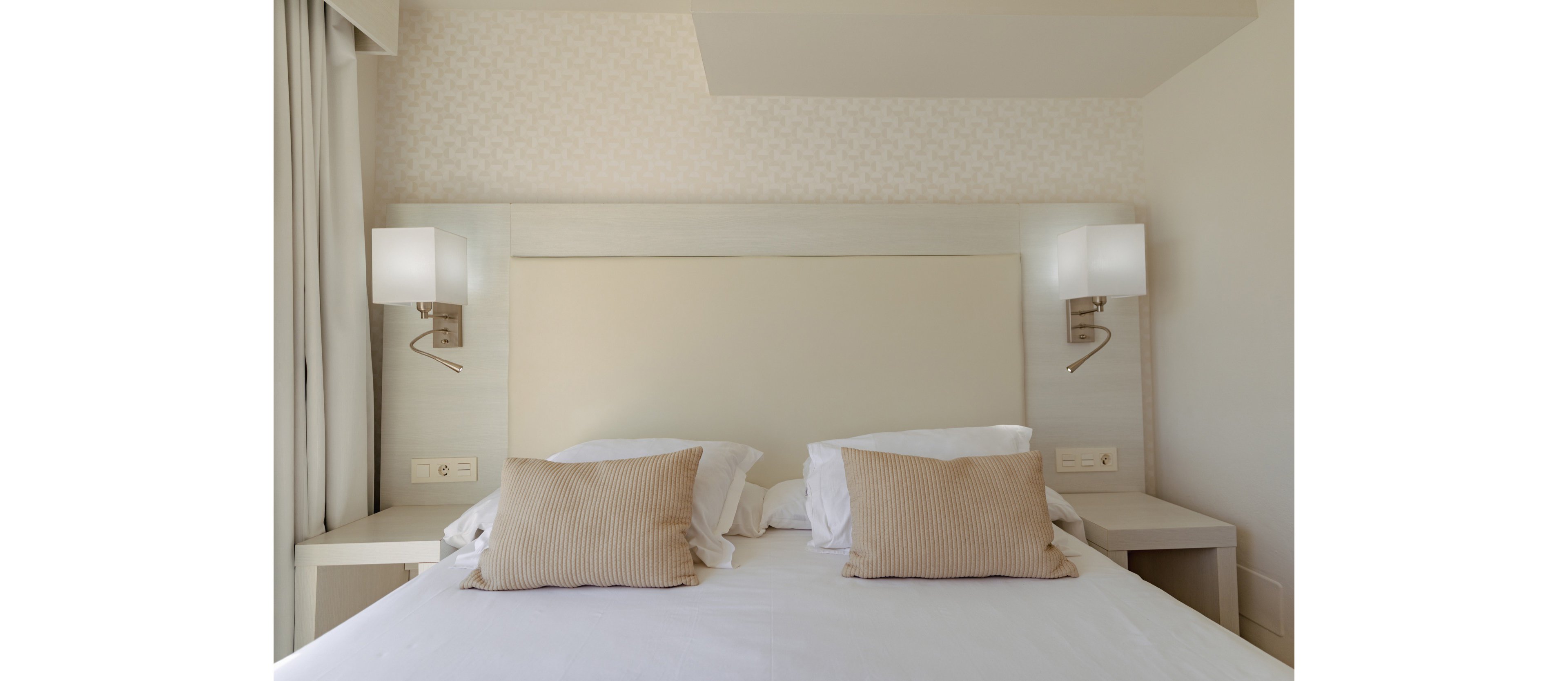 A neatly made double bed with two beige striped pillows and white linens is centered in a minimalist hotel room. Wall-mounted bedside lamps flank a light-colored headboard, creating a symmetrical composition. Neutral tones and soft lighting give the space a calm, contemporary feel. The overall design emphasizes cleanliness, comfort, and modern simplicity.
