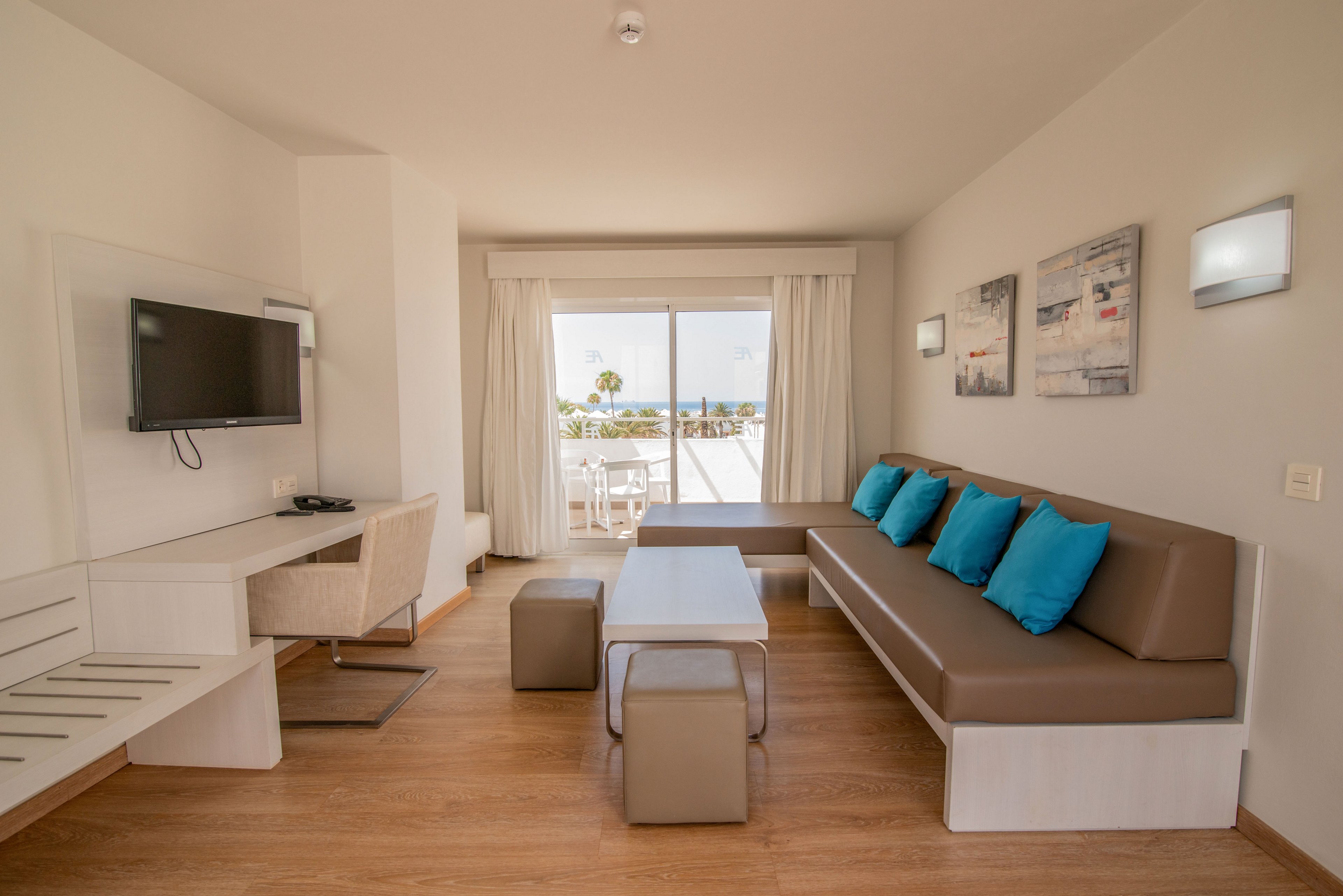 A contemporary hotel suite features a long brown sofa with bright turquoise cushions against a neutral wall. A flat-screen television is mounted opposite a minimalist desk and chair. Light wood flooring and white furniture create an airy, clean atmosphere. Large sliding glass doors open to a balcony with outdoor seating and a distant view of palm trees and water.