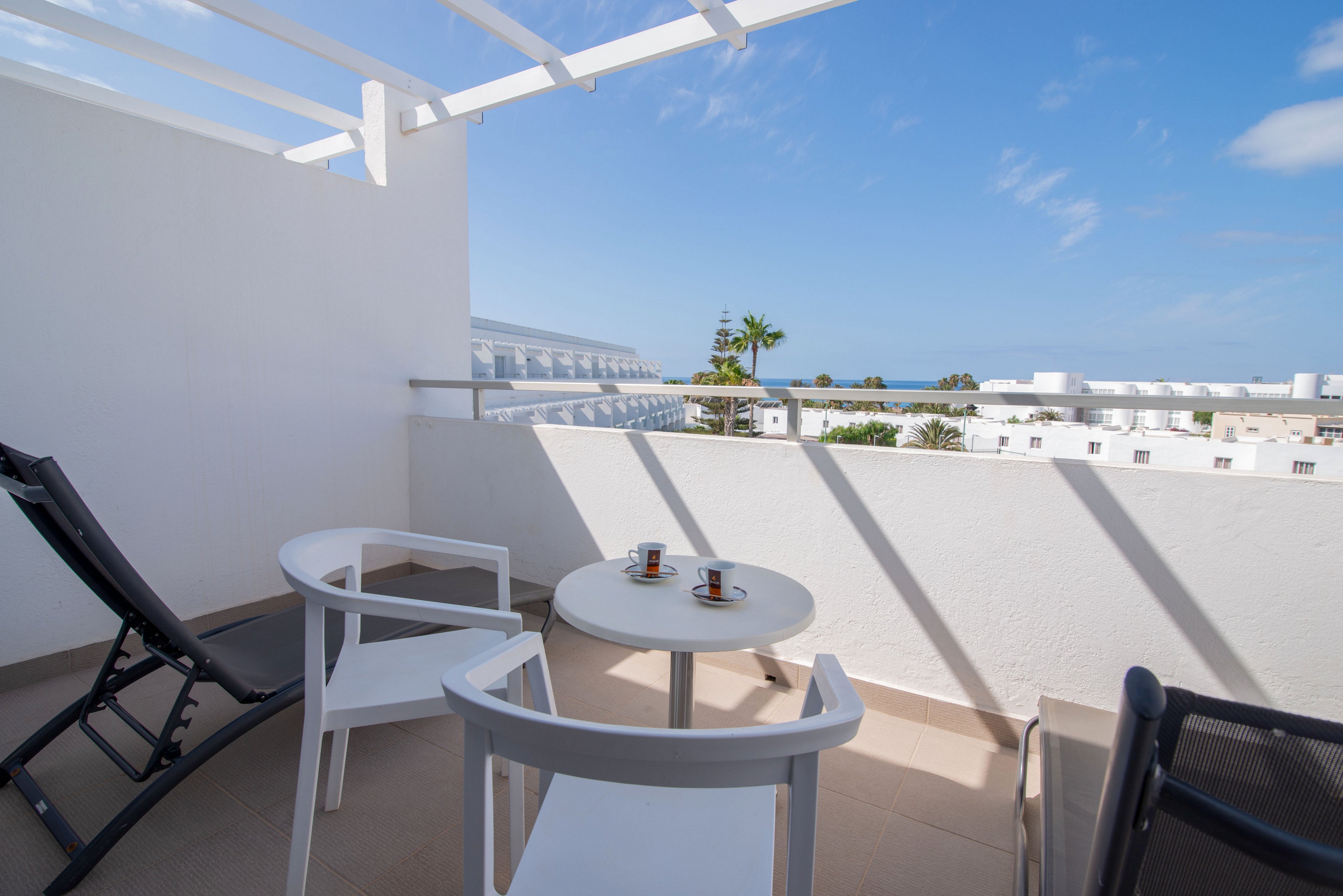 A bright hotel balcony features a small round table with two white chairs and a black lounge chair. Two espresso cups sit on the table, suggesting a relaxing coffee break. The terrace overlooks white buildings, palm trees, and a distant view of the sea under a clear blue sky. Strong sunlight casts sharp shadows, emphasizing the clean, modern design of the outdoor space.