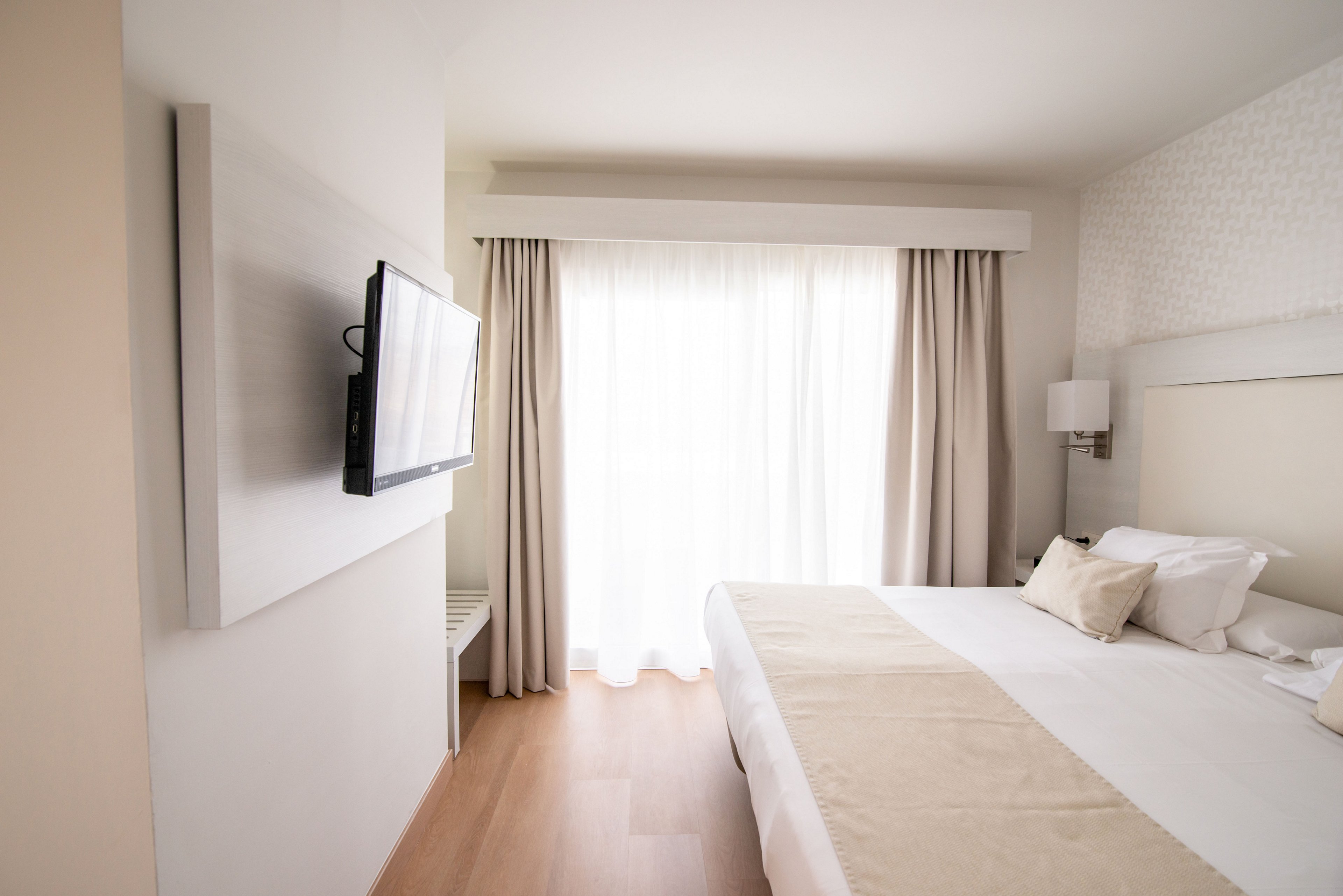 A modern hotel bedroom features a neatly made double bed with neutral-colored linens and pillows. A flat-screen television is mounted on the wall opposite the bed. Sheer curtains and heavier drapes cover a large window or balcony door, allowing soft natural light to fill the room. The decor is minimalist with light wood flooring and white walls, creating a clean and relaxing atmosphere.