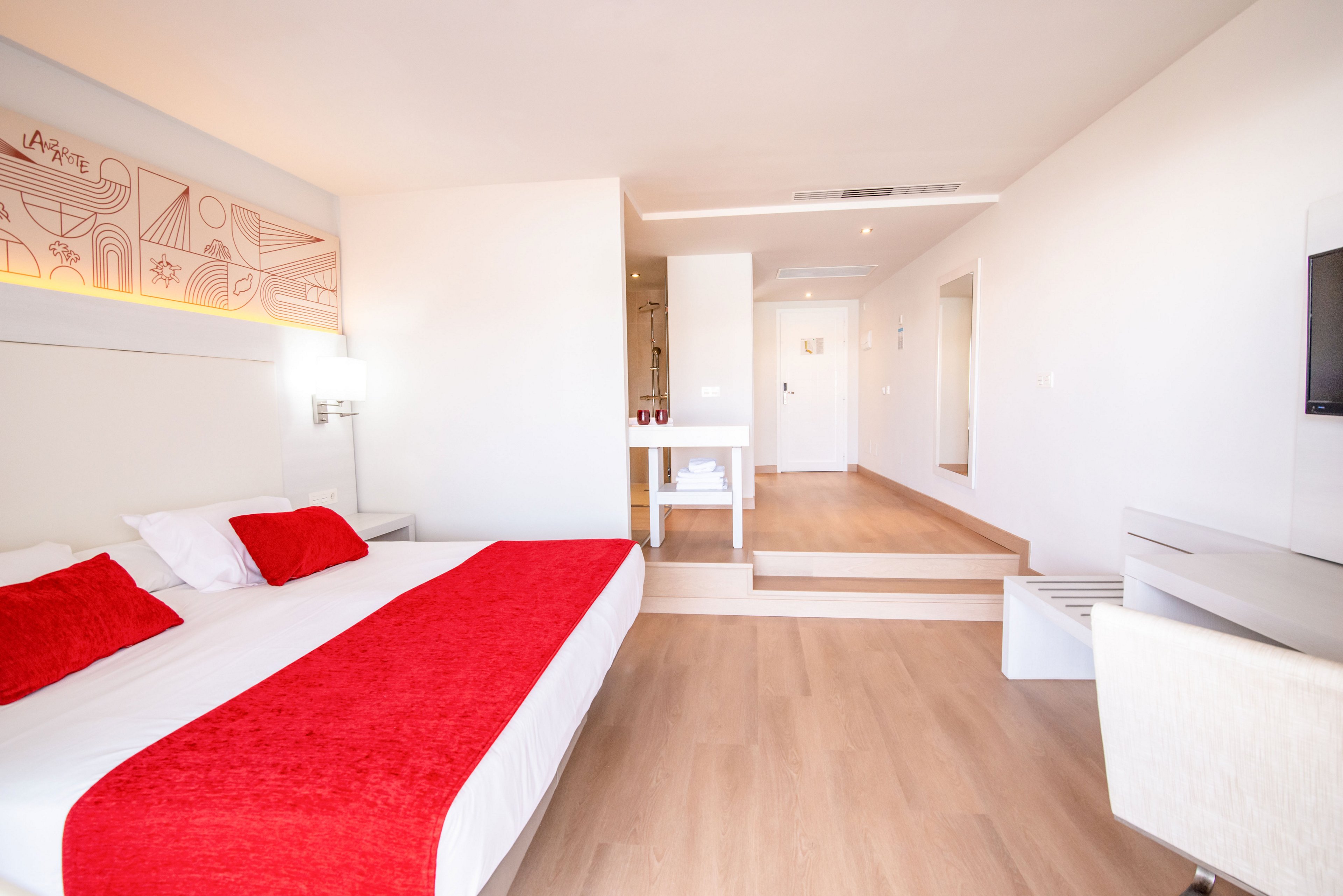 Spacious contemporary hotel room featuring a large bed with white linens and vivid red pillows and throw. The interior is decorated in a minimalist style with light wood flooring and white walls. A small sitting or work area with a chair and desk faces a wall-mounted television. Soft lighting and clean lines create an airy, inviting atmosphere.