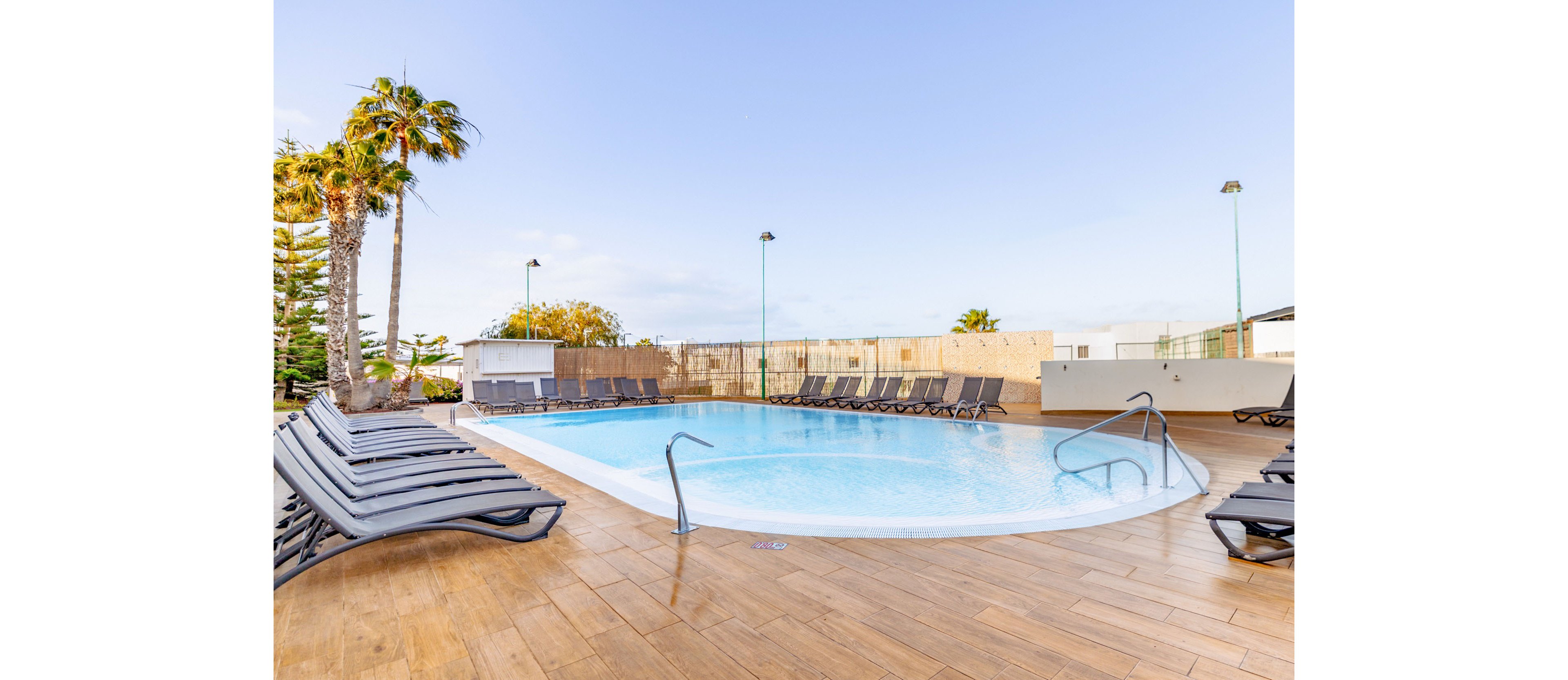 A clear blue swimming pool sits at the center of an outdoor resort area. Numerous dark sun loungers surround the pool on a wooden deck, all unoccupied. Palm trees and other greenery rise behind a fence, with low white buildings in the background. The sky is bright and clear, suggesting a calm, sunny day at the pool.