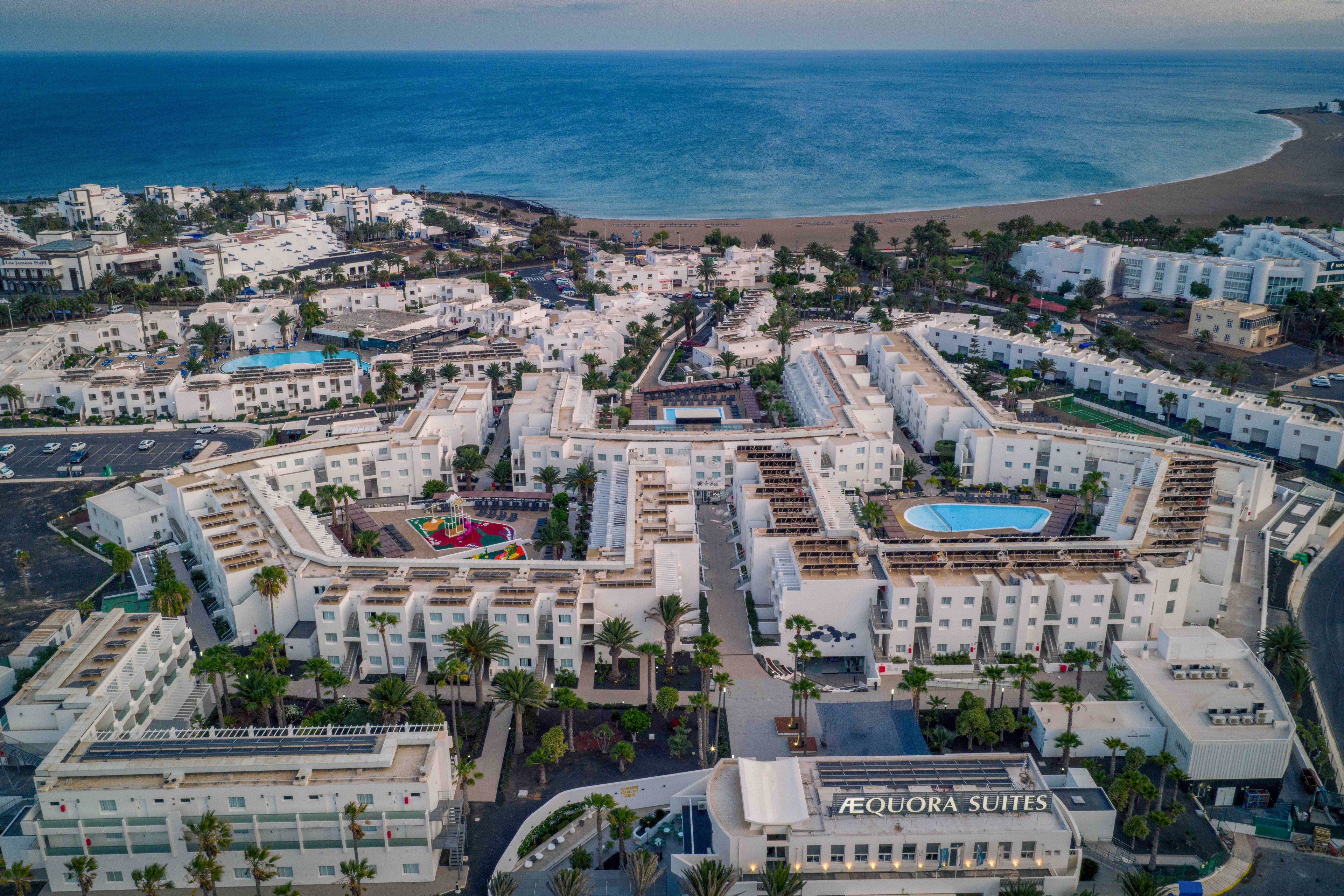 A large white resort complex is captured from above near a coastline with calm blue water. Multiple buildings with flat roofs surround a central courtyard and a bright blue swimming pool. Palm trees and landscaped paths run through the property, with a small colorful playground visible among the structures. The surrounding area includes additional hotels and residential buildings stretching toward the beach and sea.