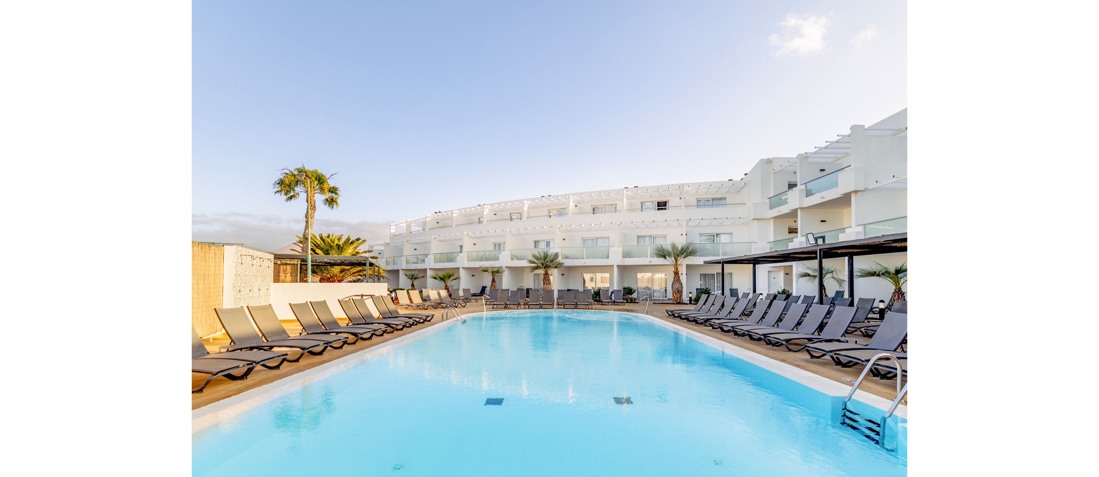 A large outdoor swimming pool is surrounded by rows of empty sun loungers at a modern white resort. The multi-story building features balconies and glass railings overlooking the pool area. Palm trees and minimal landscaping add a tropical touch to the clean, contemporary setting. The bright daylight and clear sky create a relaxed vacation atmosphere.