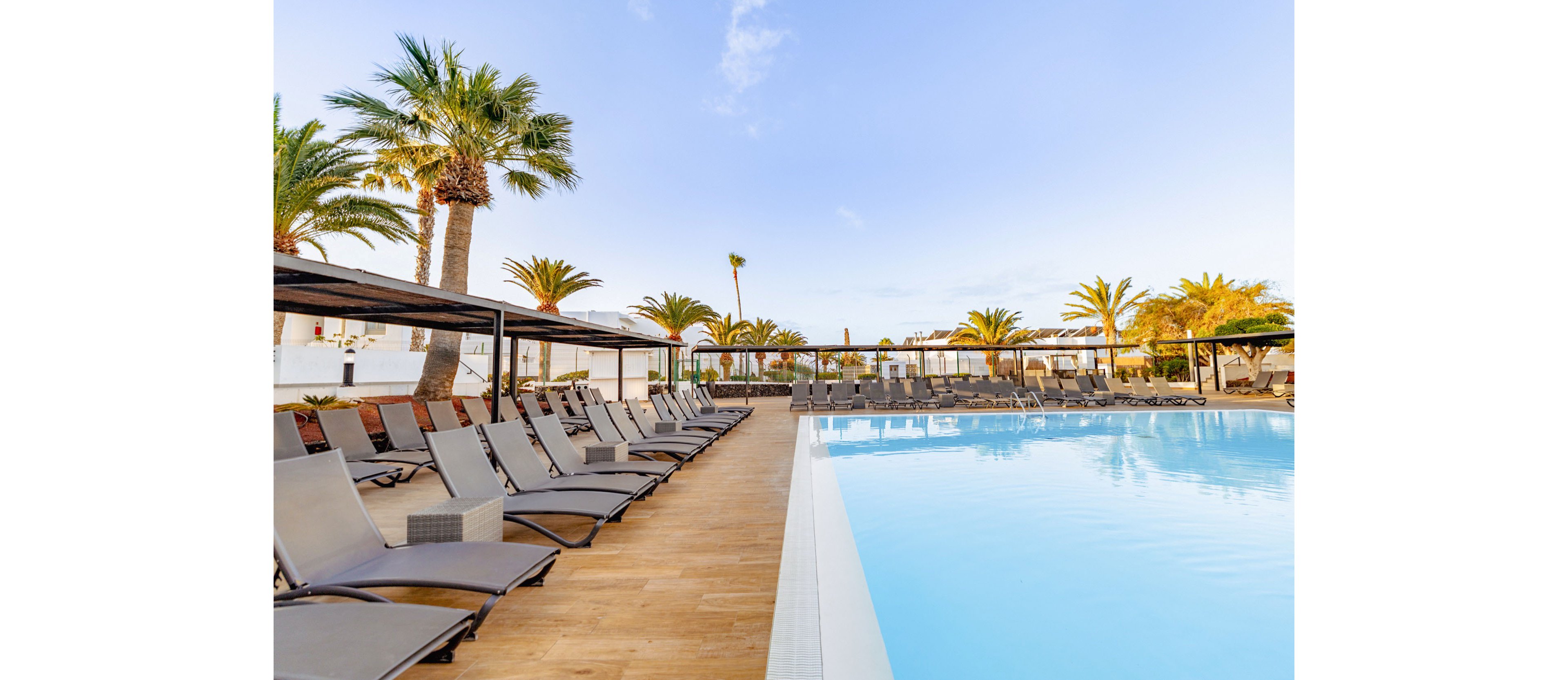 A large outdoor swimming pool sits beside rows of empty sun loungers at a resort. The scene features wooden decking, shaded pergolas, and numerous reclining chairs arranged neatly along the poolside. Tall palm trees and white buildings frame the background under a clear blue sky. The bright daylight and calm water create a relaxed, vacation atmosphere.