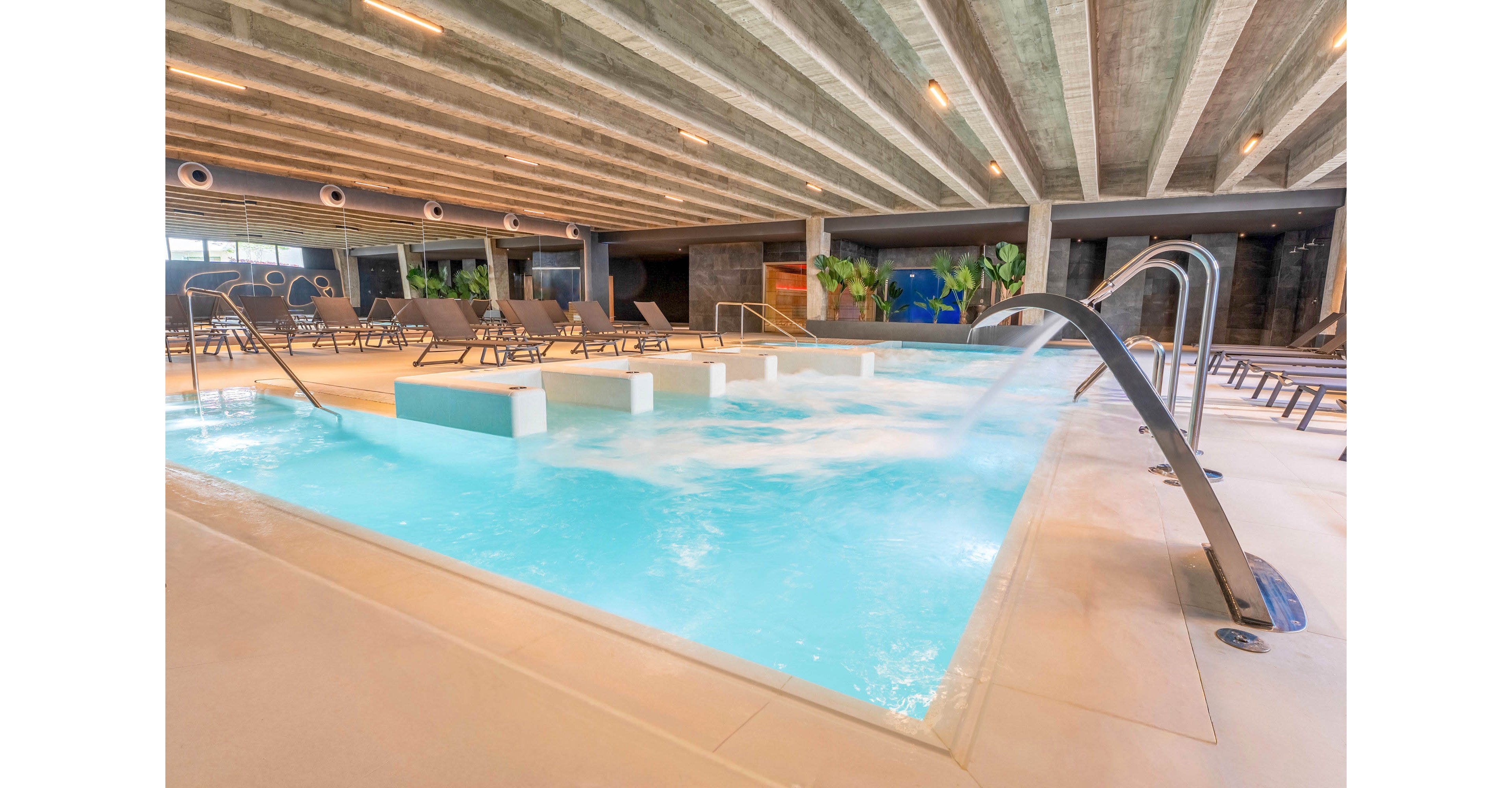 A contemporary indoor spa pool features powerful hydrotherapy jets and bubbling water. The pool is surrounded by lounge chairs and a smooth light-colored deck under an exposed concrete ceiling. Curved metal handrails and water features create a sleek, modern look. In the background, plants and warm lighting add a relaxing, resort-like atmosphere.