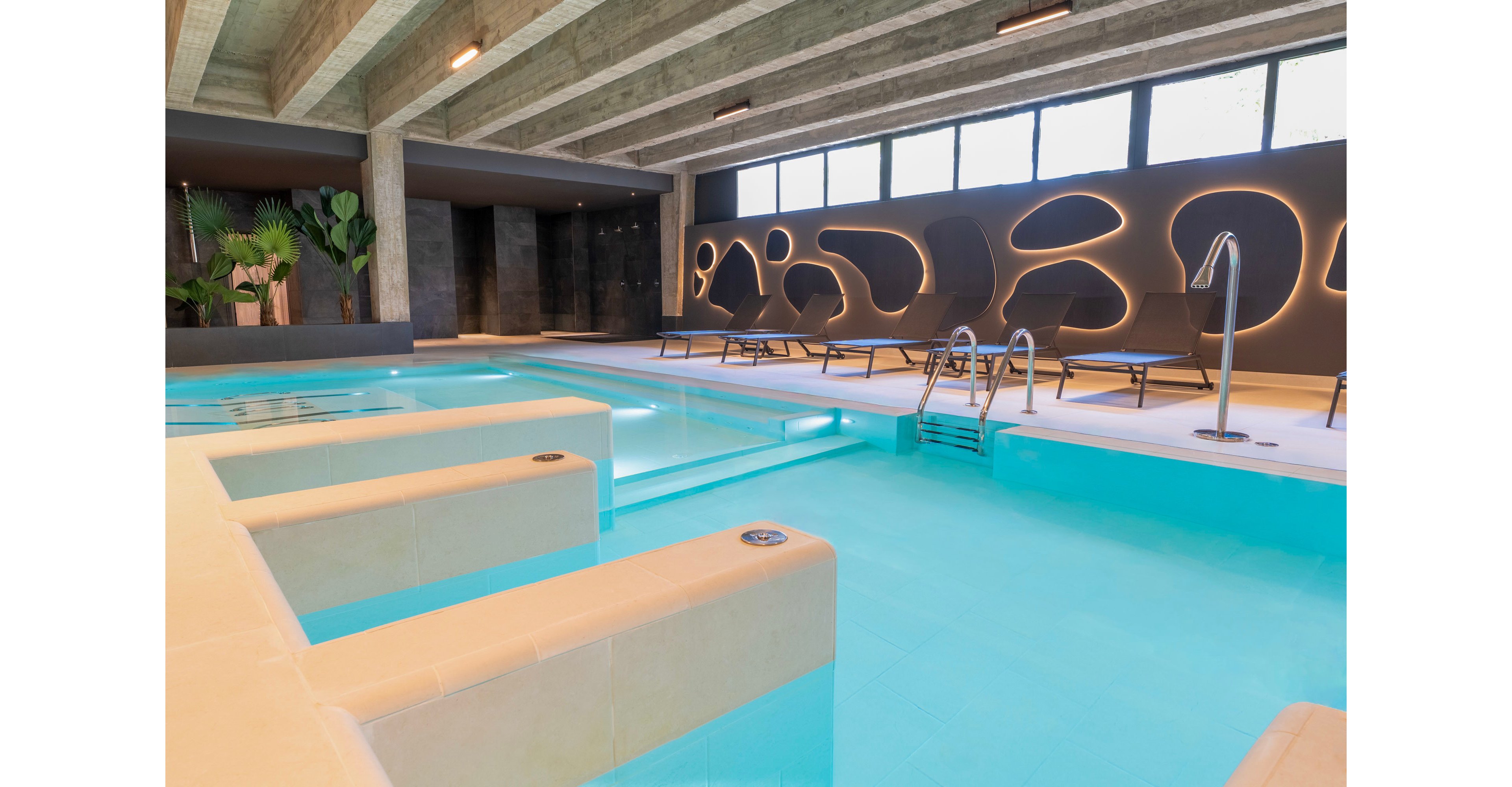 A contemporary indoor spa pool features clear blue water and built-in seating areas. The space is framed by exposed concrete beams and large windows that let in natural light. Sculptural wall panels with integrated lighting create a stylish backdrop behind several lounge chairs. The overall atmosphere is calm and luxurious, ideal for relaxation and wellness.