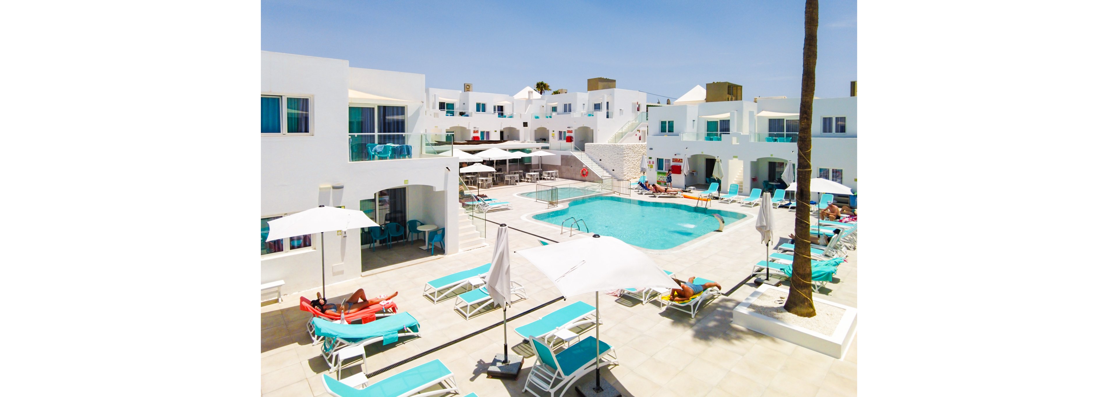 A vibrant resort featuring a central swimming pool surrounded by white modern buildings. Lounge chairs and umbrellas create a relaxing atmosphere under a bright blue sky. Guests are seen enjoying the sunny day, adding a lively touch to the serene setting.