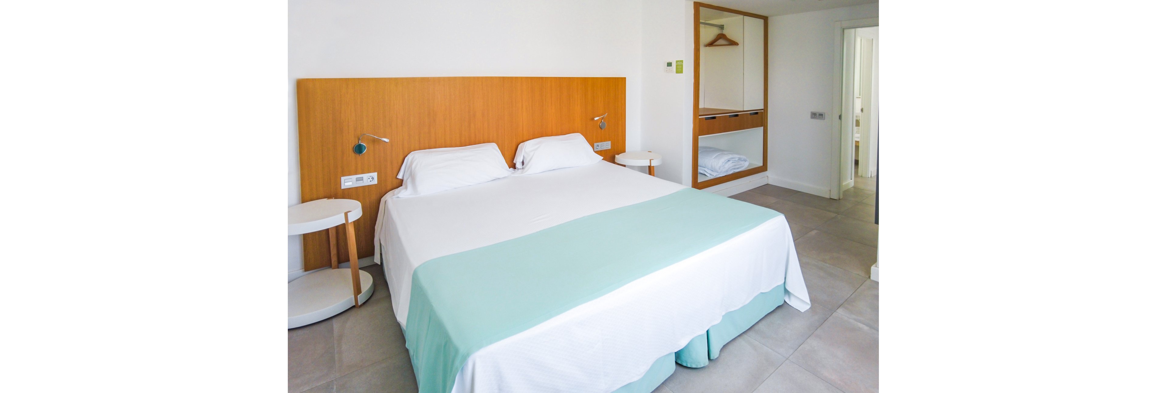 A clean and minimalist hotel room featuring a double bed with white linens and a light turquoise bed runner. The setting includes wooden furniture, bedside tables, and an open wardrobe with hangers. The room is brightly lit with natural light and has a neutral color palette.