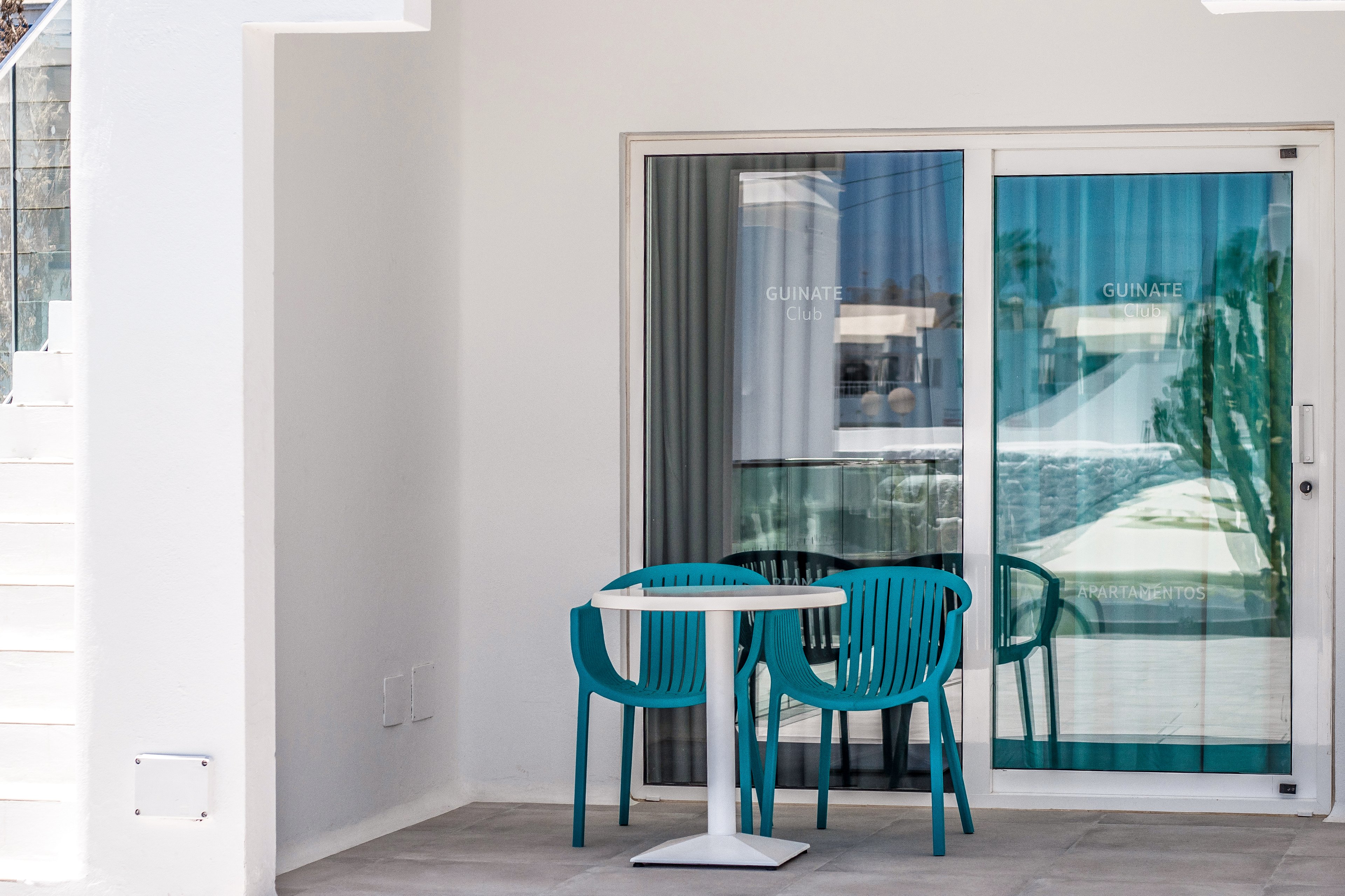 A minimalist outdoor patio featuring a white table and two turquoise chairs. The setting includes a glass sliding door with visible text 'GUINAZ' and a reflection of a pool area. The clean, modern design emphasizes a relaxed and bright atmosphere.