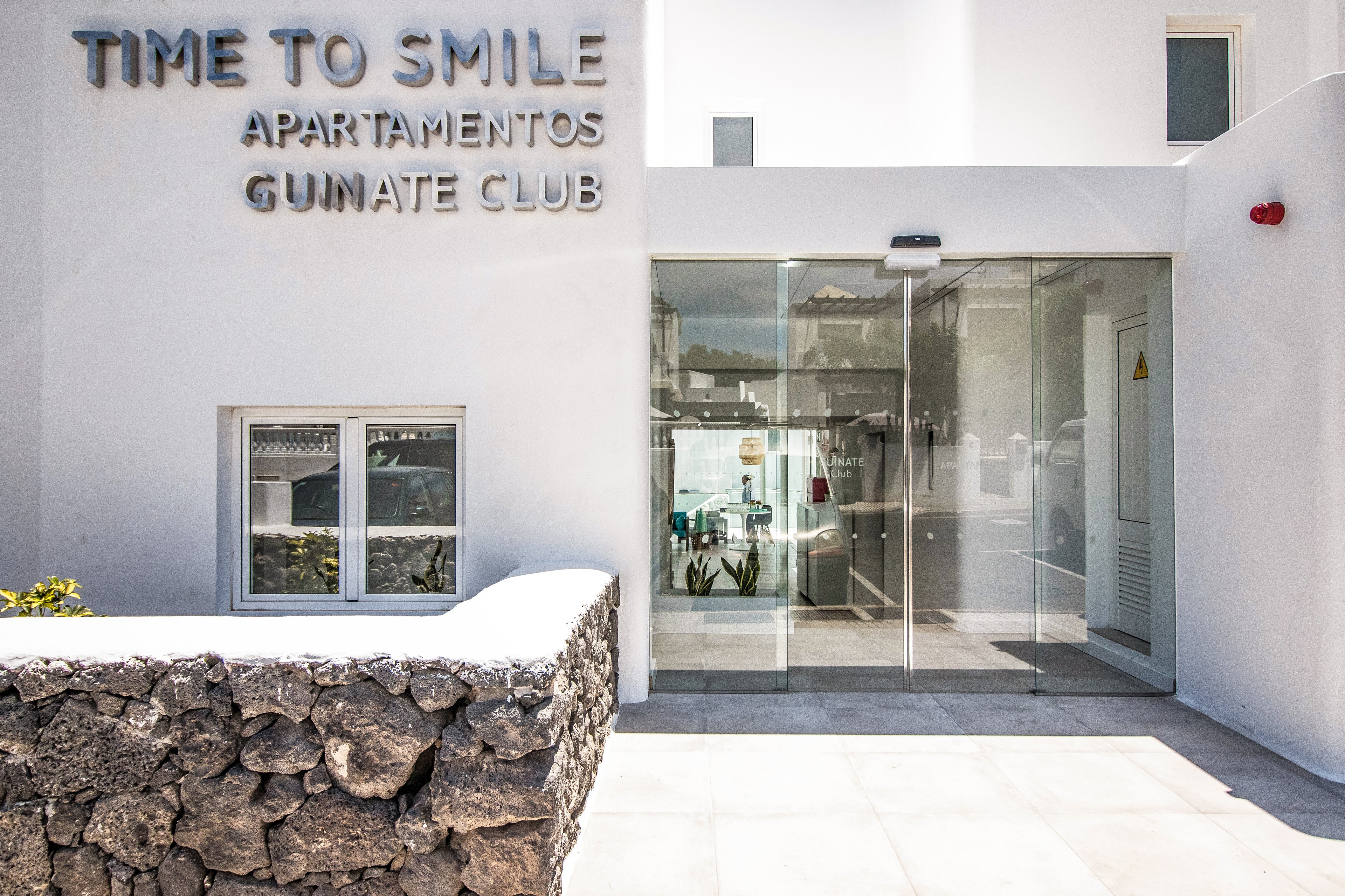 The image showcases the entrance of 'Time to Smile Apartamentos Guinate Club,' featuring clean, modern architecture with white walls and glass doors. A stone wall adds texture to the foreground, while the signage is prominently displayed on the building facade. The setting appears bright and sunny, suggesting a vacation or leisure environment.