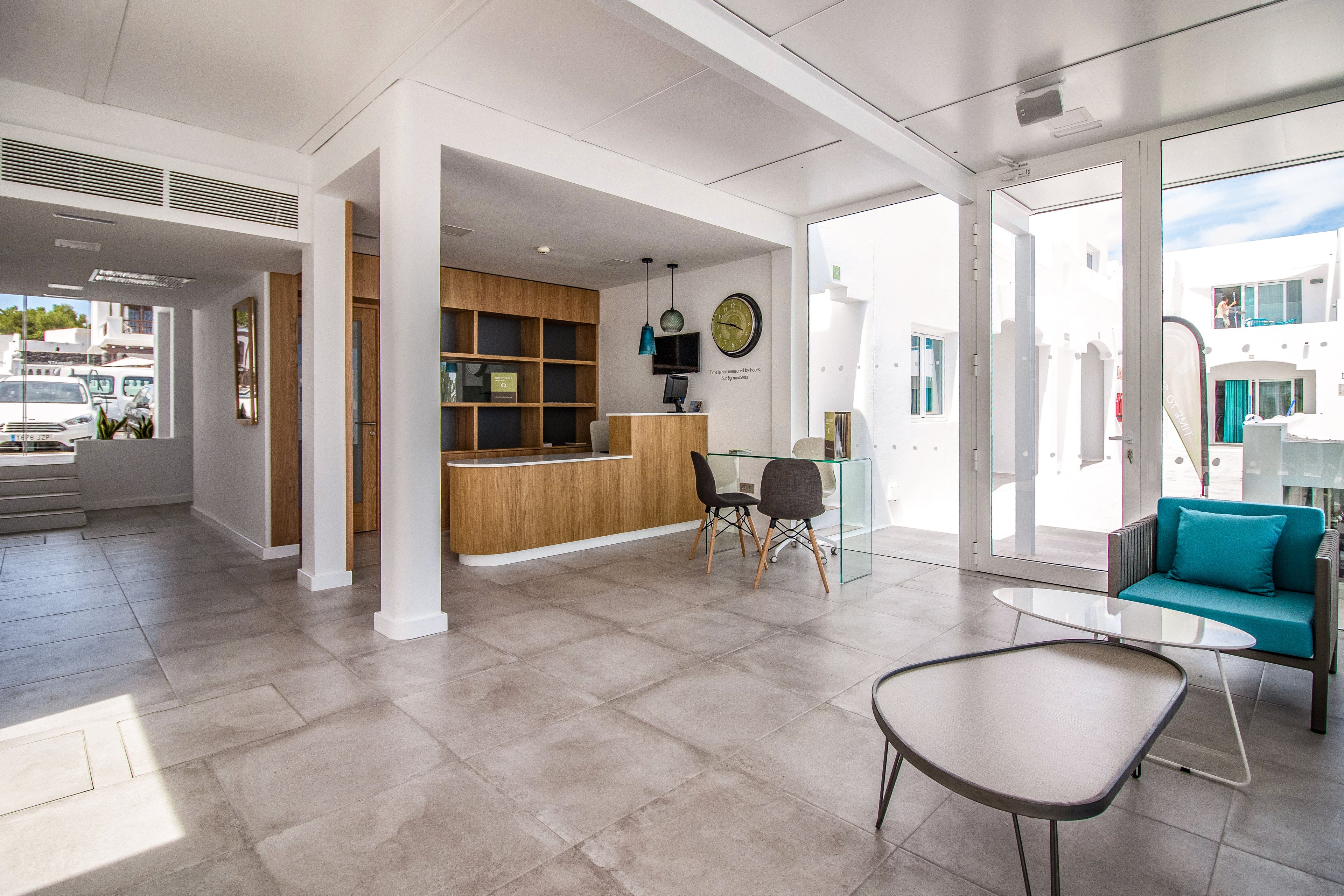 A contemporary reception area featuring minimalist furniture and ample natural light. The space includes a wooden desk, glass table, and teal-accented chairs, creating a welcoming atmosphere. Large windows provide views of a bright outdoor setting with white buildings. The design emphasizes clean lines and a neutral palette with pops of color.