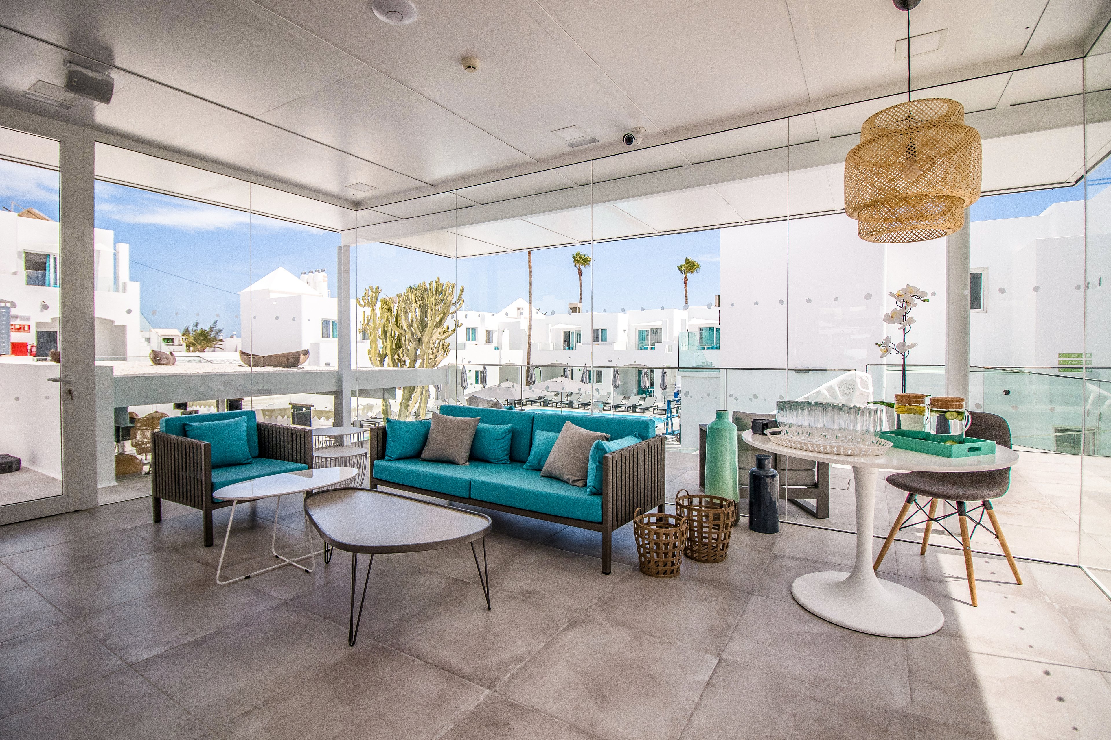 A stylish outdoor seating area featuring turquoise cushions on a modern sofa and chairs. The setting is a bright, open terrace surrounded by white buildings and clear glass panels. Decorative elements include woven baskets, a hanging lamp, and a sleek white dining table. The scene is bathed in natural light, creating a fresh and inviting atmosphere.