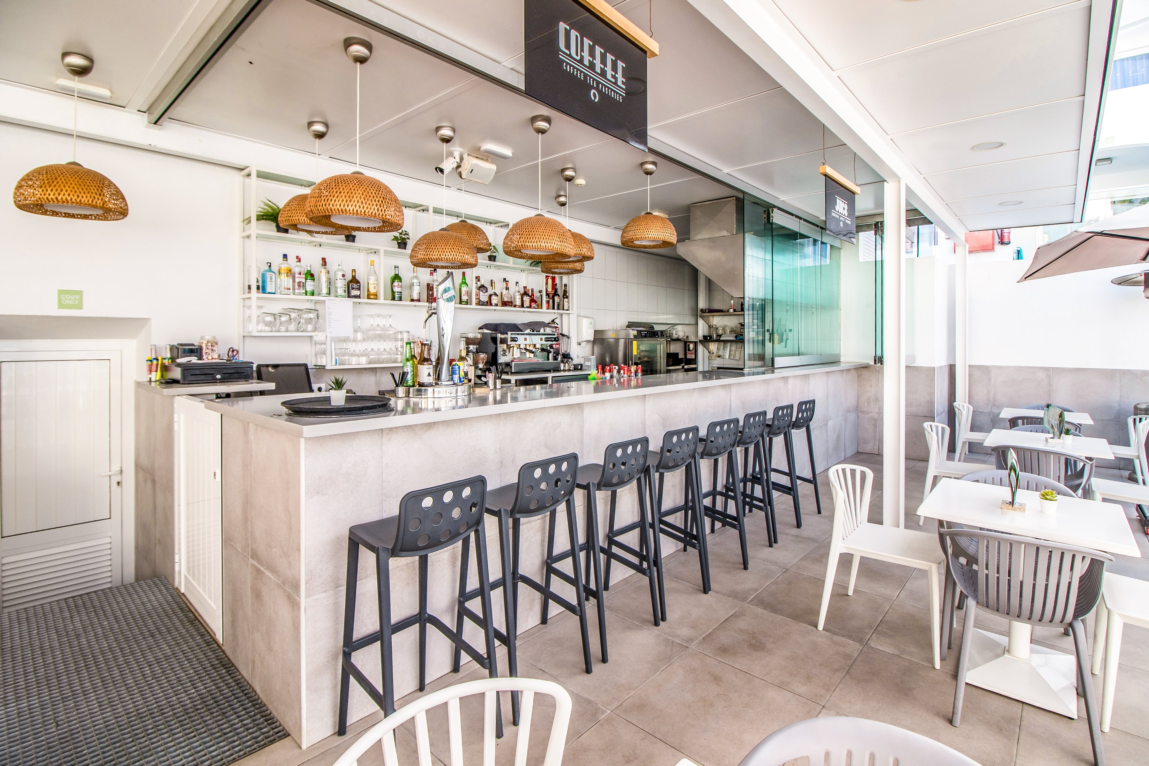 A contemporary coffee bar featuring a sleek counter with black stools and wicker pendant lights. The setting includes a bright, open space with white tables and chairs, creating a casual and inviting atmosphere. Visible signage reads 'COFFEE' above the counter, emphasizing the establishment's focus. Bottles and coffee-making equipment are displayed behind the counter.