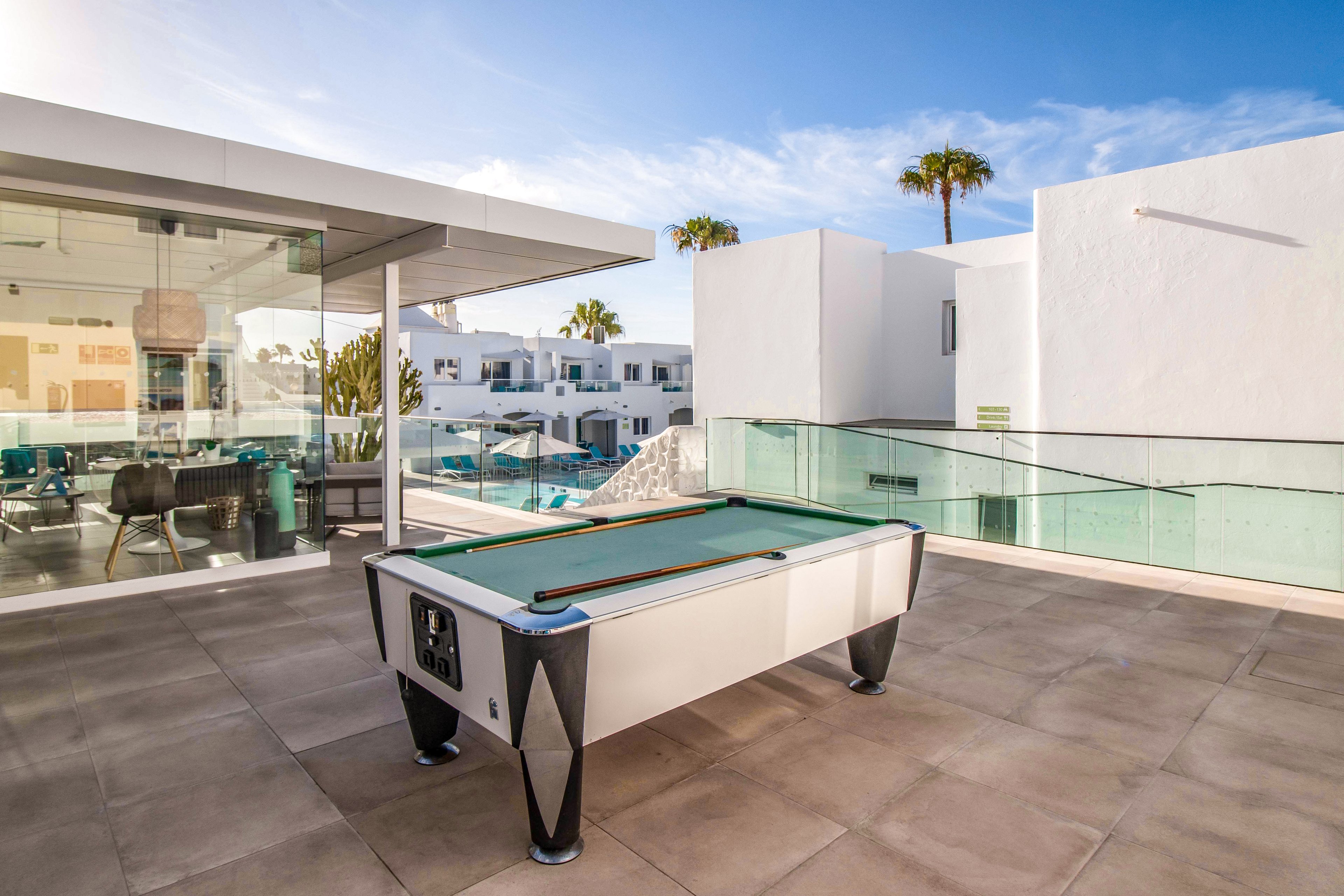 A sleek outdoor pool table is set on a spacious modern terrace with glass railings. The setting features white minimalist architecture, a clear blue sky, and palm trees in the background. The scene exudes a relaxed and luxurious vibe, perfect for leisure activities.