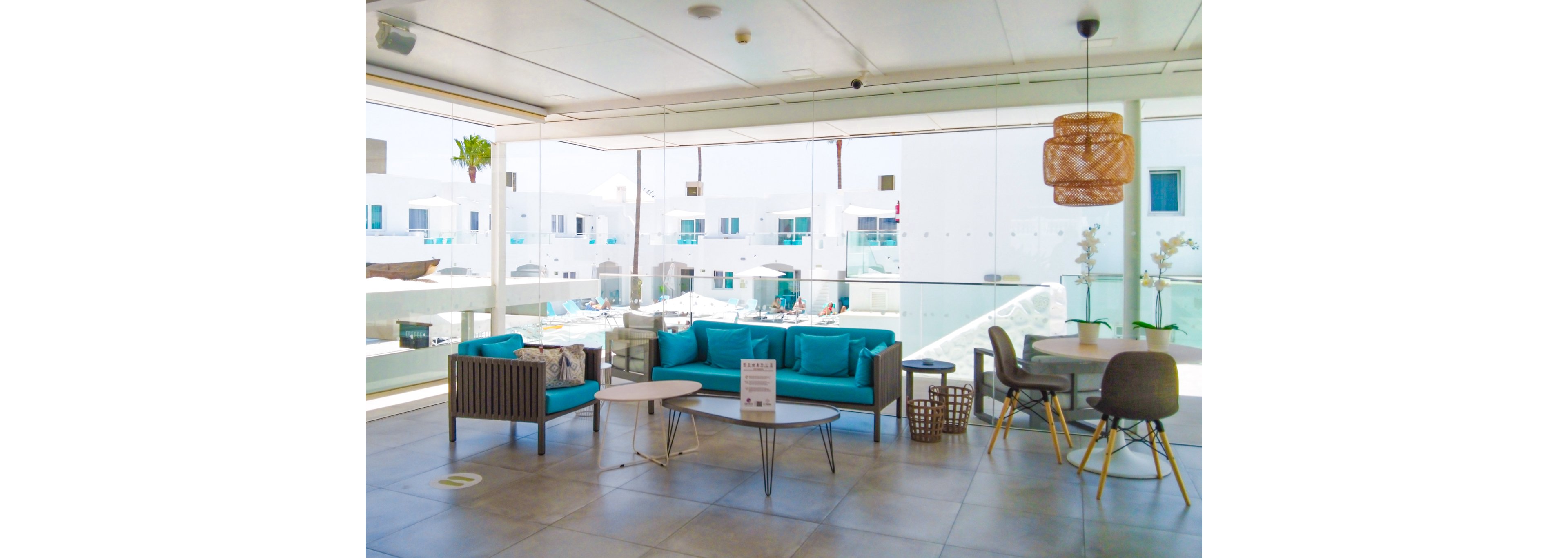 A contemporary indoor lounge area featuring turquoise cushions on wooden furniture, complemented by a glass wall overlooking a bright outdoor space. The setting includes a mix of seating options, pendant lighting, and decorative plants. Visible in the background are white buildings and a poolside area with umbrellas.