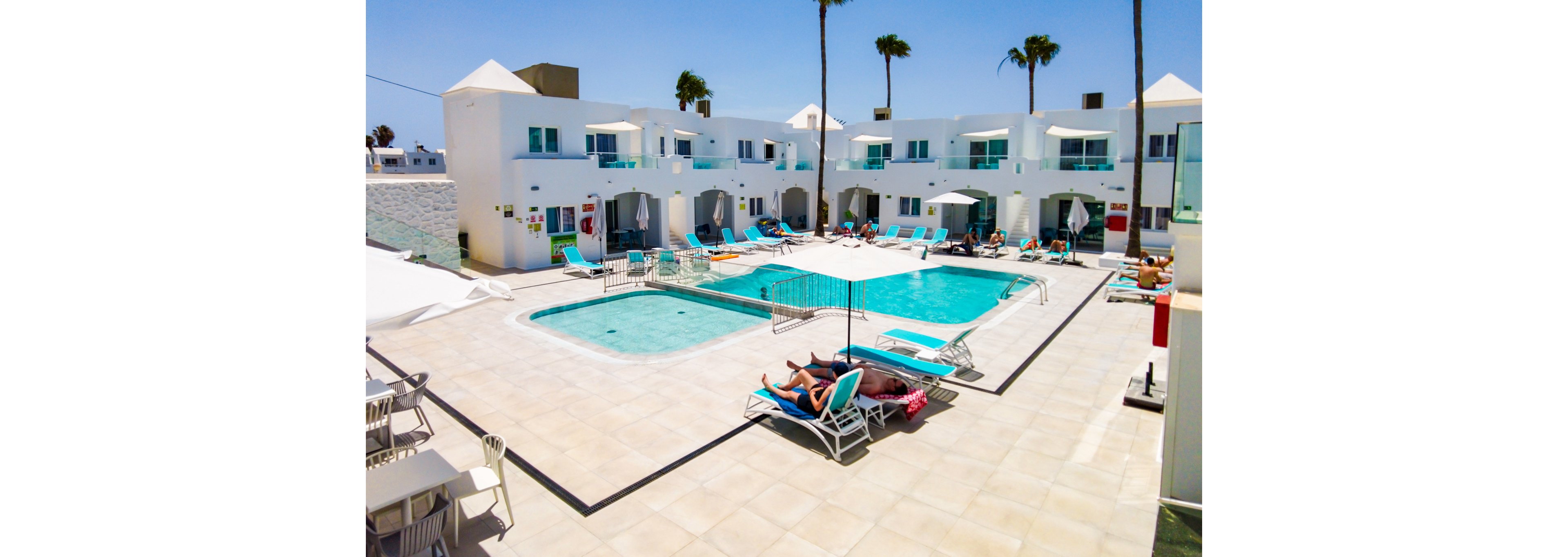 A bright and modern resort featuring a central swimming pool surrounded by white buildings and palm trees. Guests relax on lounge chairs under the sun, with umbrellas providing shade. The setting is clean and inviting, evoking a tropical vacation atmosphere. The clear blue sky enhances the vibrant and serene mood.