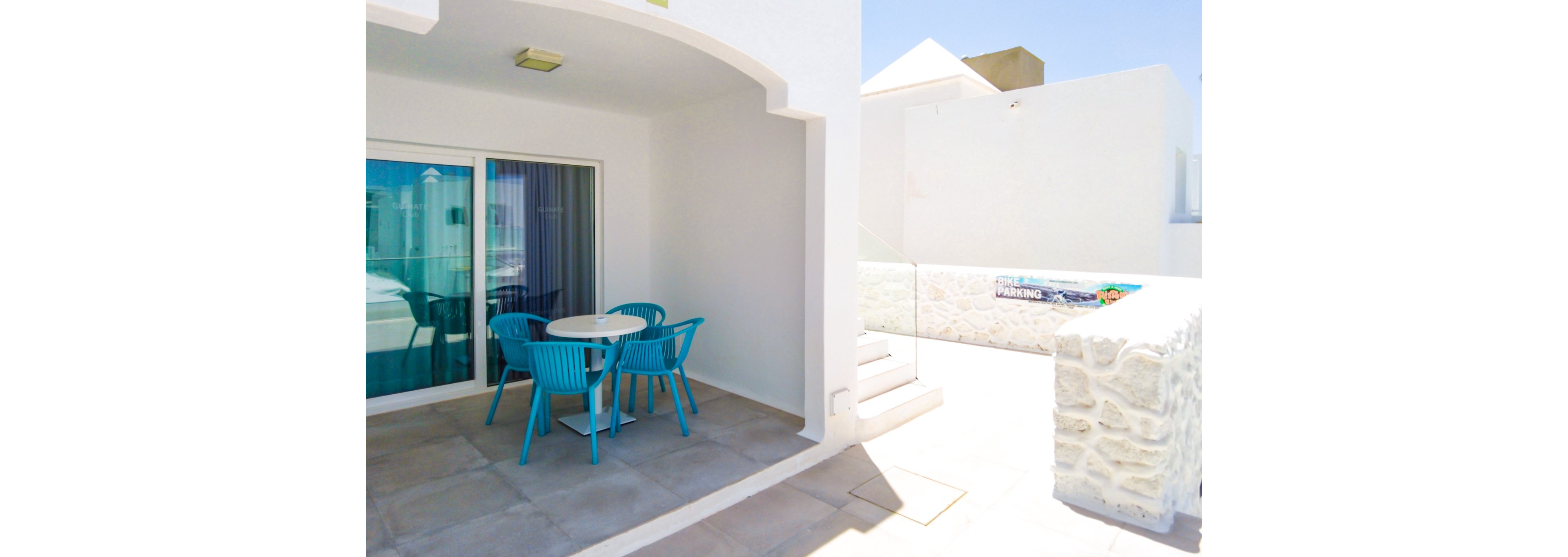 A bright and minimalist outdoor patio featuring a white-walled setting with a small table and four vibrant blue chairs. The area is partially shaded and opens to a sunny space with a glass barrier. Visible text includes 'PARKING' on a nearby sign, adding a functional detail to the scene.
