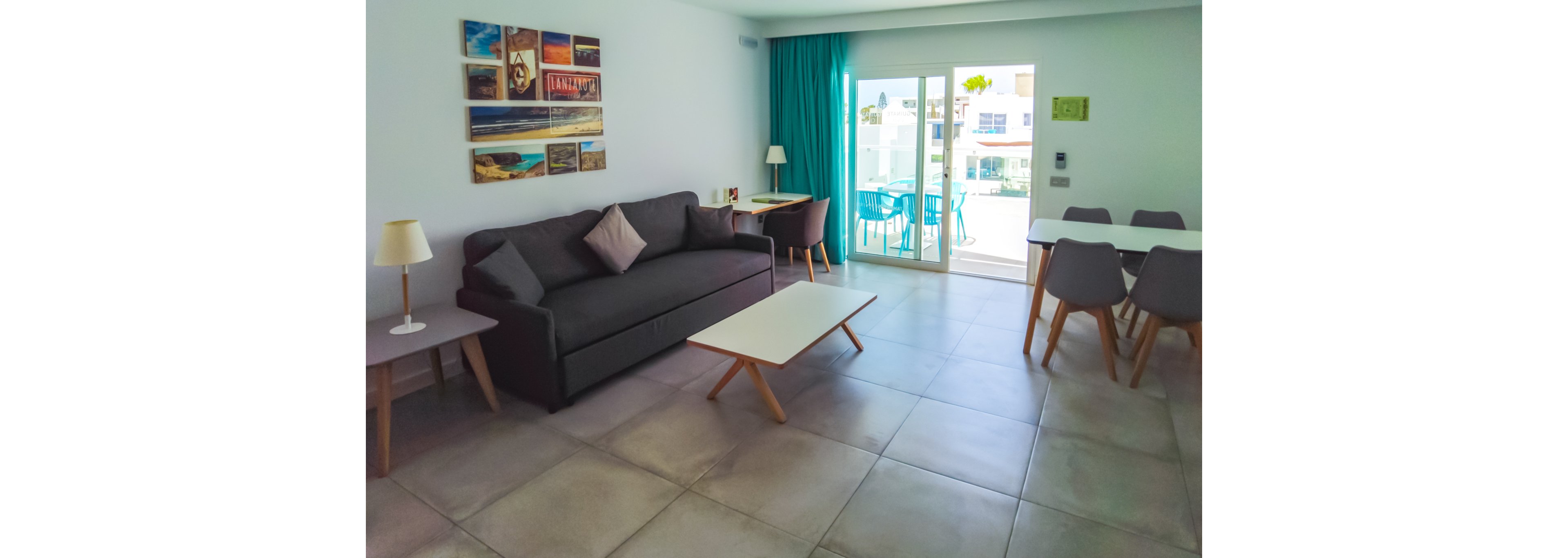 A bright and modern living room featuring a gray sofa, white coffee table, and dining area with gray chairs. The space is illuminated by natural light from a sliding glass door leading to a balcony with blue chairs. Wall art and minimalist decor enhance the clean, contemporary style.