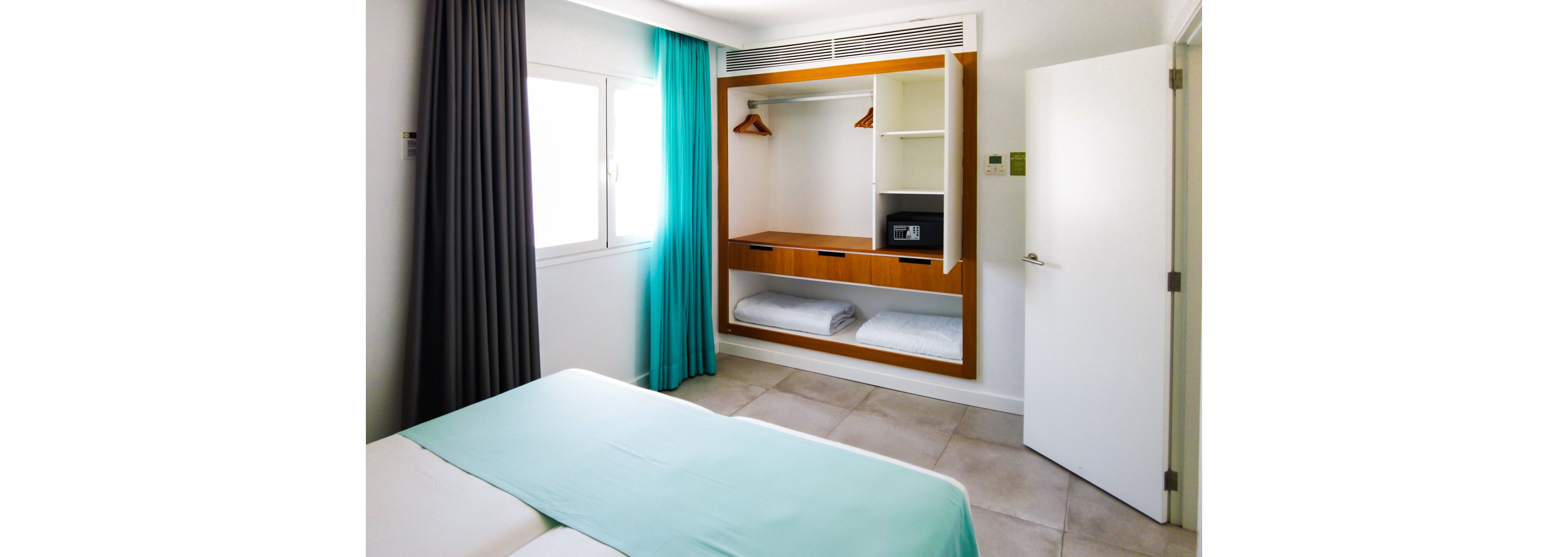 A bright and minimalist hotel room featuring an open wardrobe with wooden accents. The setting includes a neatly made bed with turquoise bedding and curtains, complemented by natural light streaming through the window. A small safe is visible inside the wardrobe, along with folded towels and hangers. The room has a clean and organized aesthetic, ideal for travelers.
