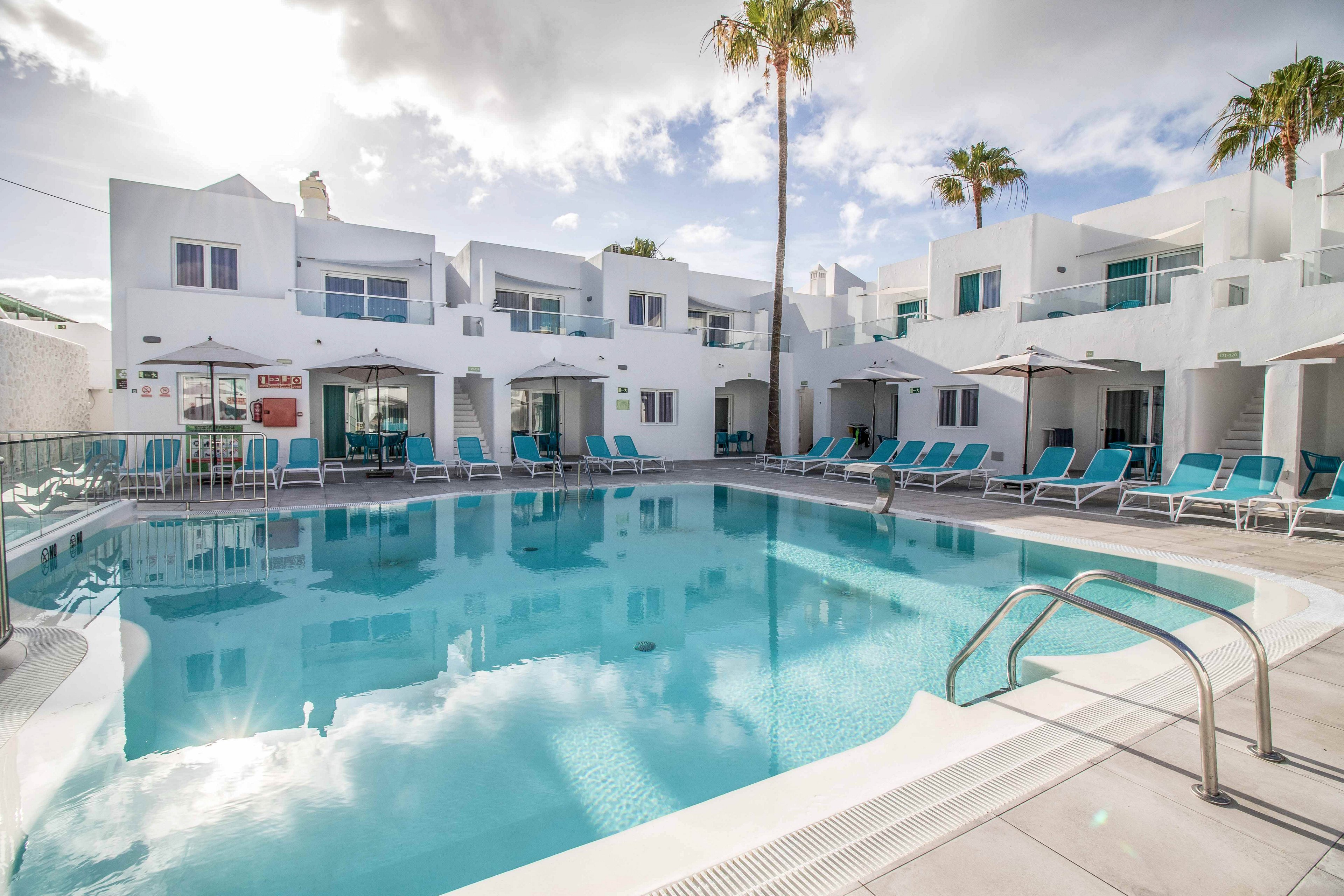 A clear blue swimming pool sits at the center of a modern white resort complex. Sun loungers with teal cushions and beige umbrellas line the poolside terrace. White two-story buildings with balconies and large windows surround the water, while tall palm trees rise above the rooftops. The bright daylight and soft clouds create a relaxed, vacation atmosphere.