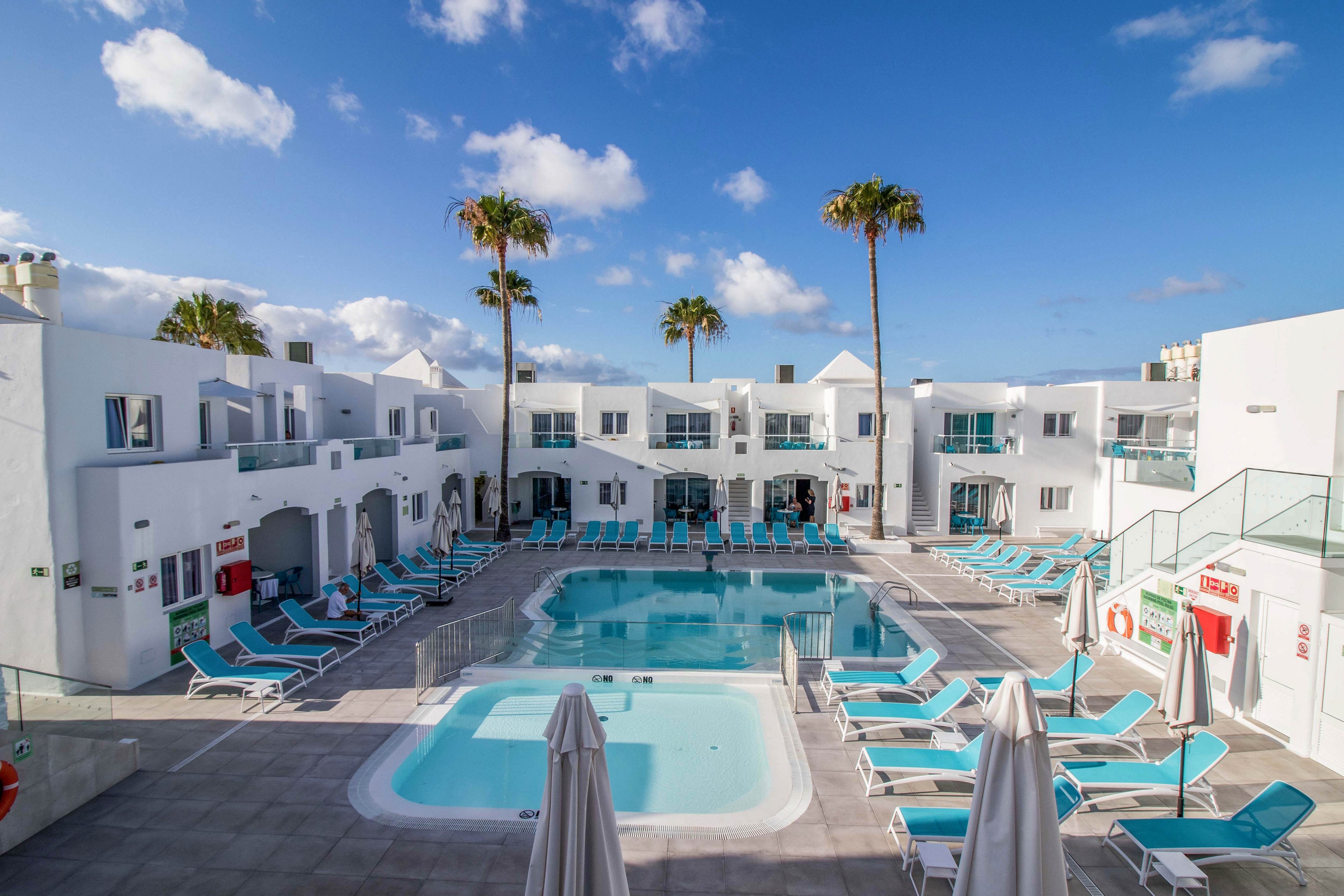A modern white resort complex surrounds a clear blue swimming pool and smaller hot tub. Numerous turquoise sun loungers and closed white umbrellas line the poolside terrace. Tall palm trees rise above the two-story buildings under a bright blue sky with scattered clouds. The scene suggests a calm, sunny holiday atmosphere at a coastal or island hotel.