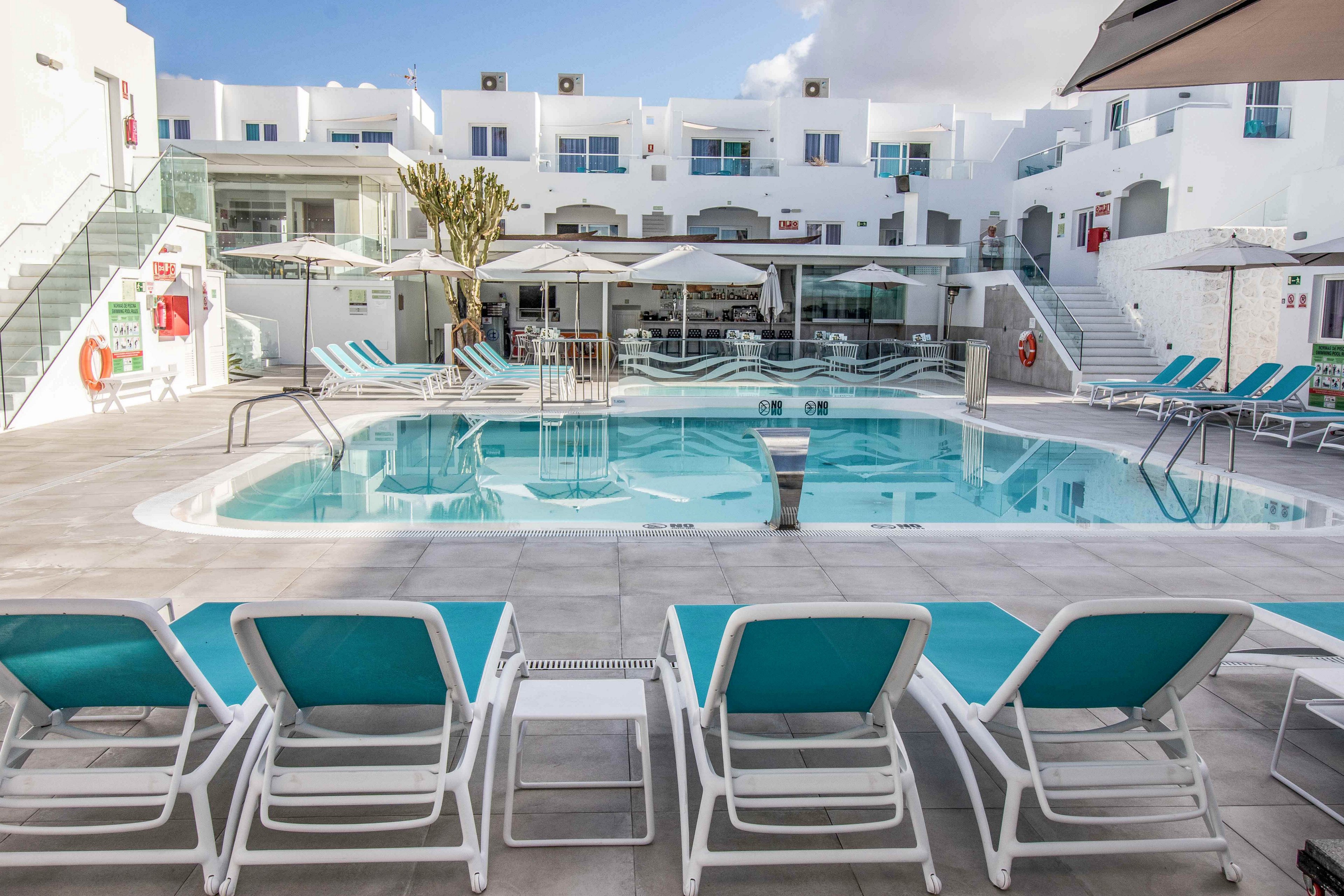 A contemporary hotel courtyard features a clear blue swimming pool surrounded by white buildings. Empty sun loungers with turquoise cushions line the poolside, creating a relaxed vacation atmosphere. Glass railings, umbrellas, and outdoor seating indicate a comfortable leisure area for guests. The bright white architecture contrasts with the blue water and sky, emphasizing a clean and modern resort setting.