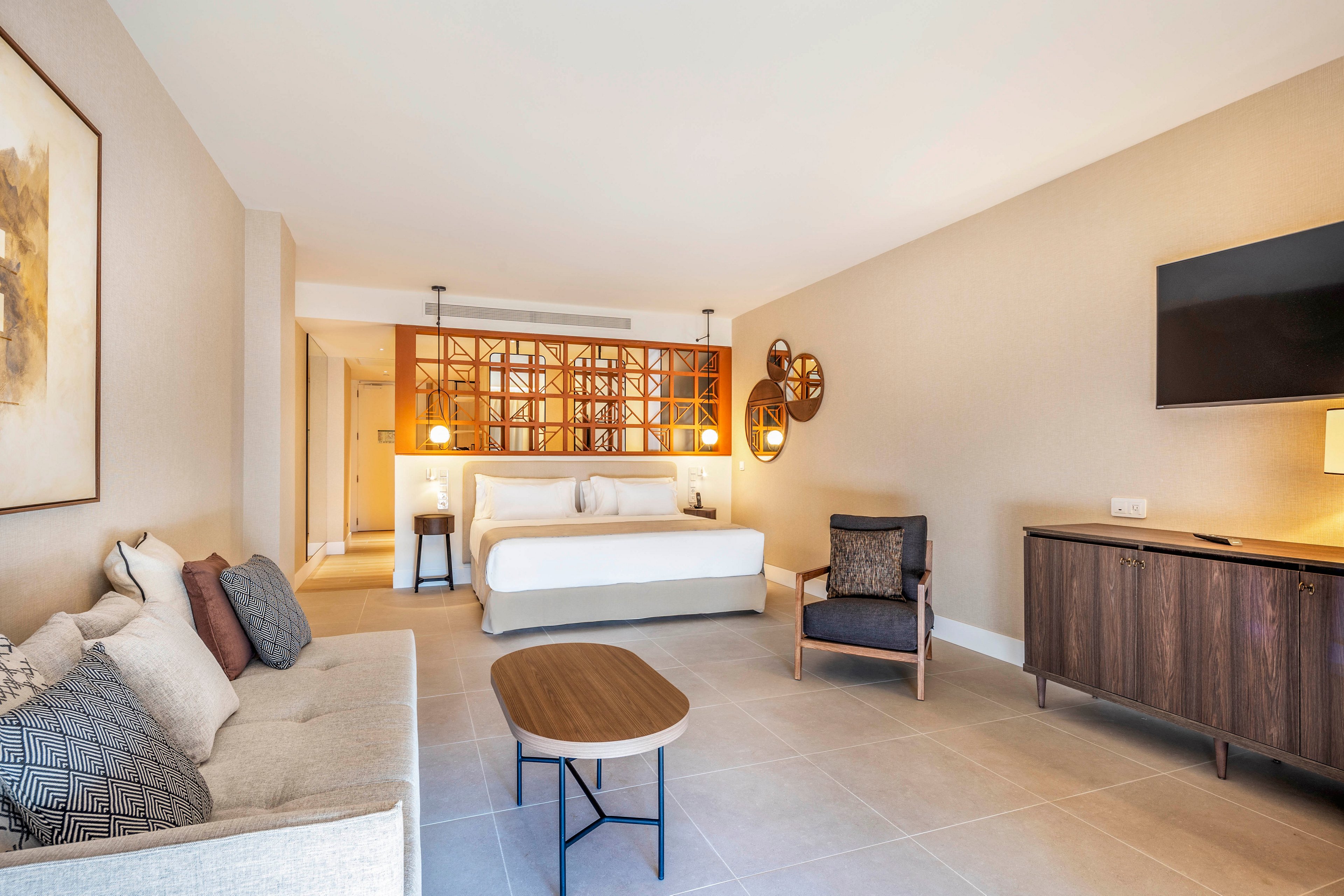 Spacious modern hotel suite featuring a large bed and cozy sitting area. The room includes a light-colored sofa with accent pillows, a wooden coffee table, and a mid-century style armchair. A wall-mounted flat-screen TV and wooden sideboard line one wall, while warm pendant and wall lamps create soft ambient lighting. Neutral tones, clean lines, and decorative wall art contribute to a calm, minimalist atmosphere.