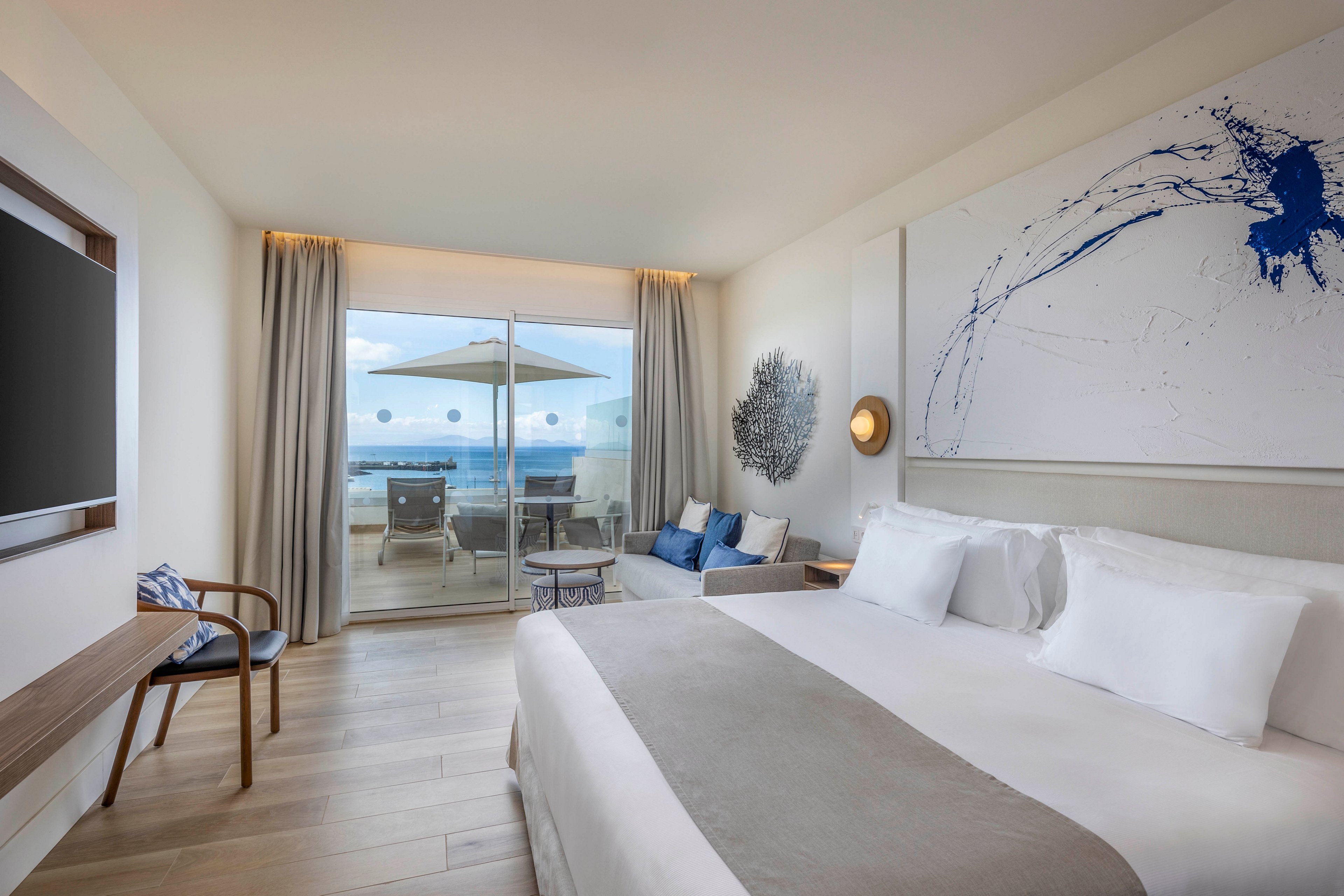 Contemporary hotel room featuring a large neatly made bed with white linens and a beige throw. The space opens onto a balcony through sliding glass doors, revealing outdoor seating and a view of the sea. Minimalist furniture, including a wooden chair, wall-mounted television, and small sofa, enhances the clean design. Neutral tones with blue accents and abstract wall art create a calm, coastal atmosphere.