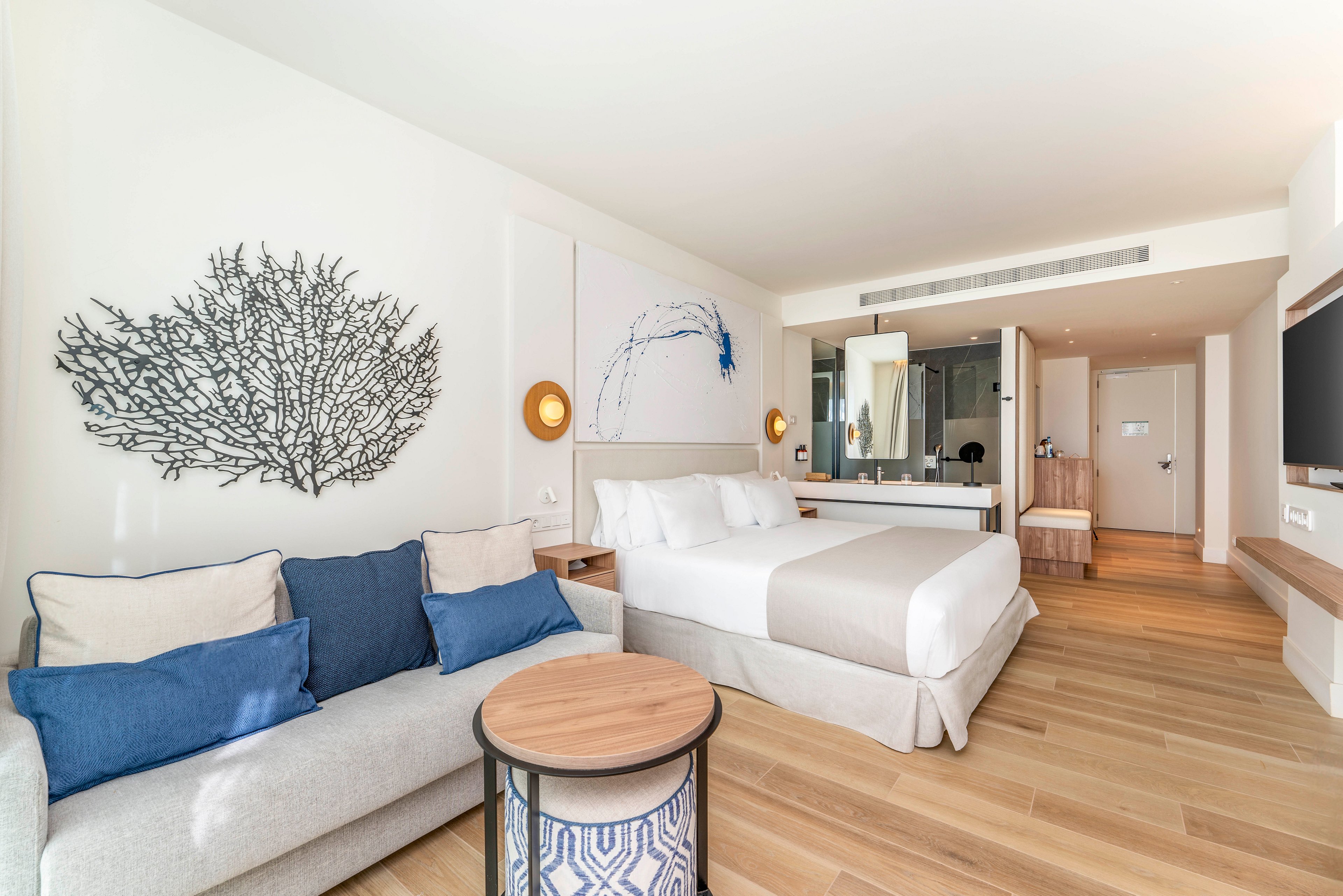A contemporary hotel room features a neatly made double bed with white linens and a beige throw. A light-colored sofa with blue accent pillows sits beside a small round wooden coffee table. The space is decorated with coastal-inspired wall art and warm wall-mounted lights. Wooden flooring, a large flat-screen TV, and an open-plan bathroom area contribute to the airy, modern atmosphere.