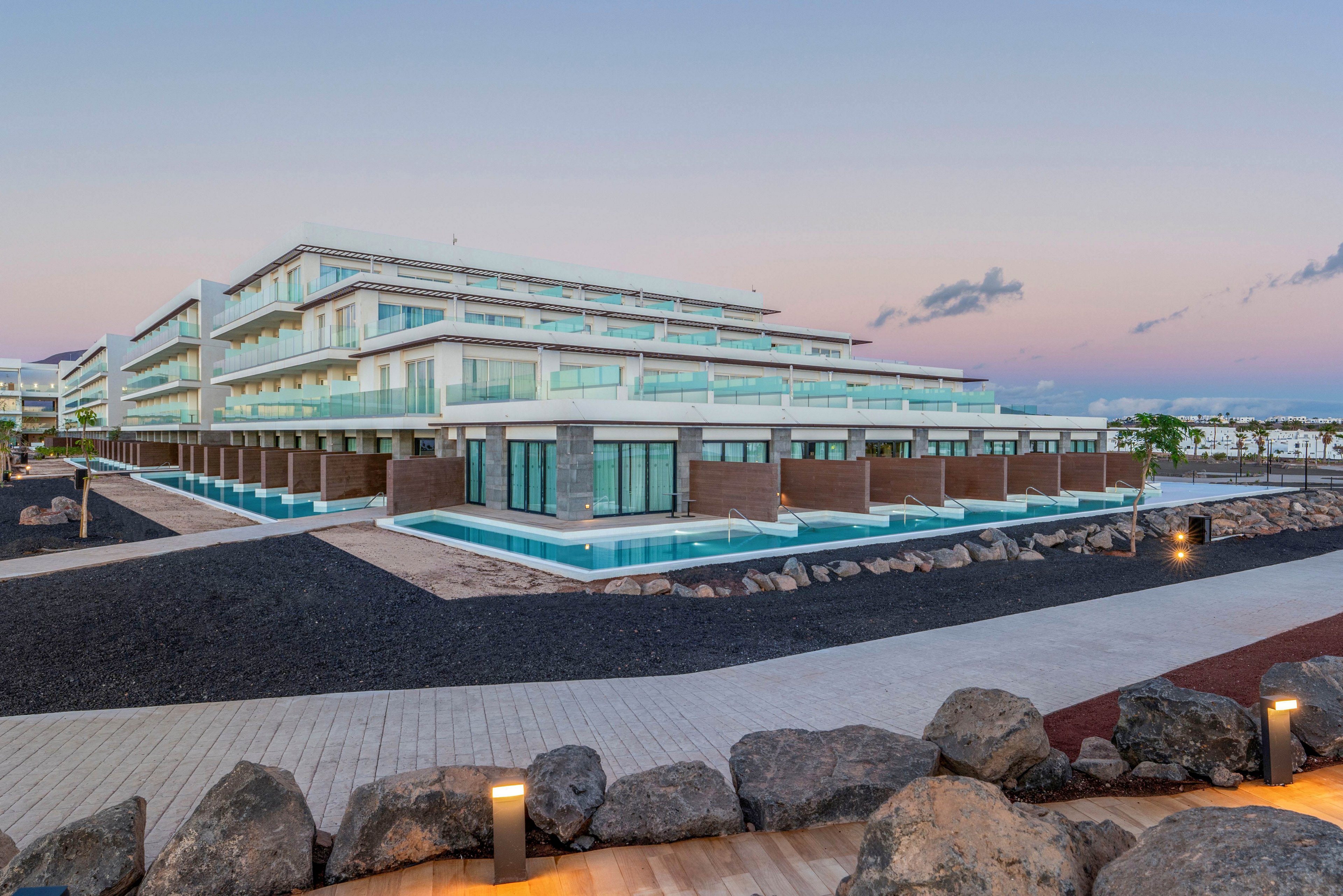 A contemporary multi-story resort hotel stands near the coastline at dusk. The building features clean lines, glass balconies, and ground-level rooms opening onto private pools. Dark volcanic rock landscaping contrasts with light wooden walkways and soft exterior lighting. The calm evening sky with pastel colors enhances the tranquil, upscale atmosphere of the property.