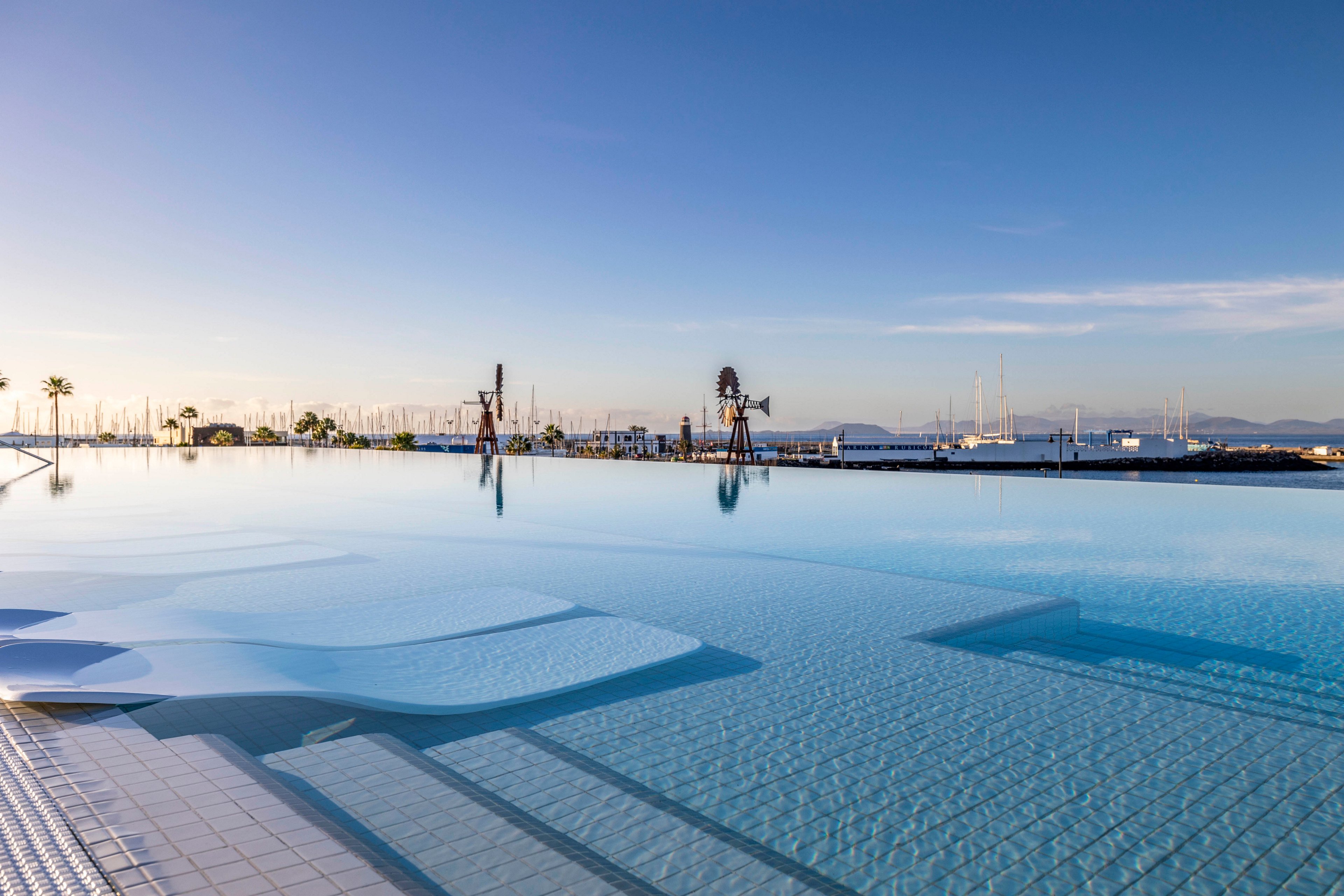 A serene infinity pool stretches toward the horizon, seamlessly blending with the calm sea. In the distance, sailboat masts rise from a marina under a clear blue sky. Minimalist white pool loungers sit partially submerged in the shallow water, emphasizing relaxation and luxury. The soft evening light creates gentle reflections on the water’s surface, enhancing the tranquil coastal atmosphere.