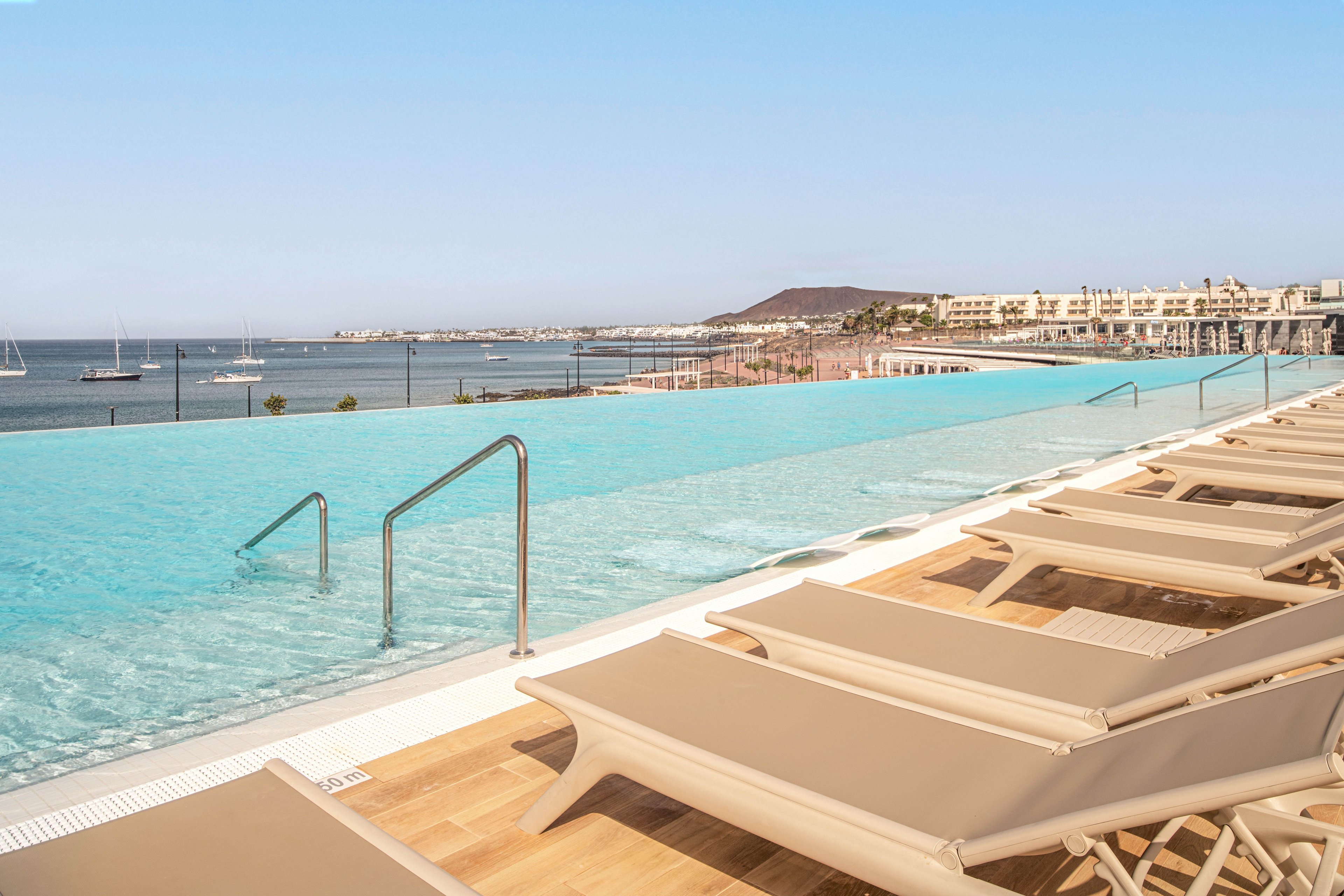 An infinity pool stretches toward a calm coastal bay, creating a seamless view between water and sea. Beige lounge chairs line the wooden deck, inviting guests to relax in the sun. In the distance, sailboats float on the water and low-rise resort buildings follow the shoreline. A hazy hill rises beyond the town under a clear blue sky, enhancing the serene vacation atmosphere.